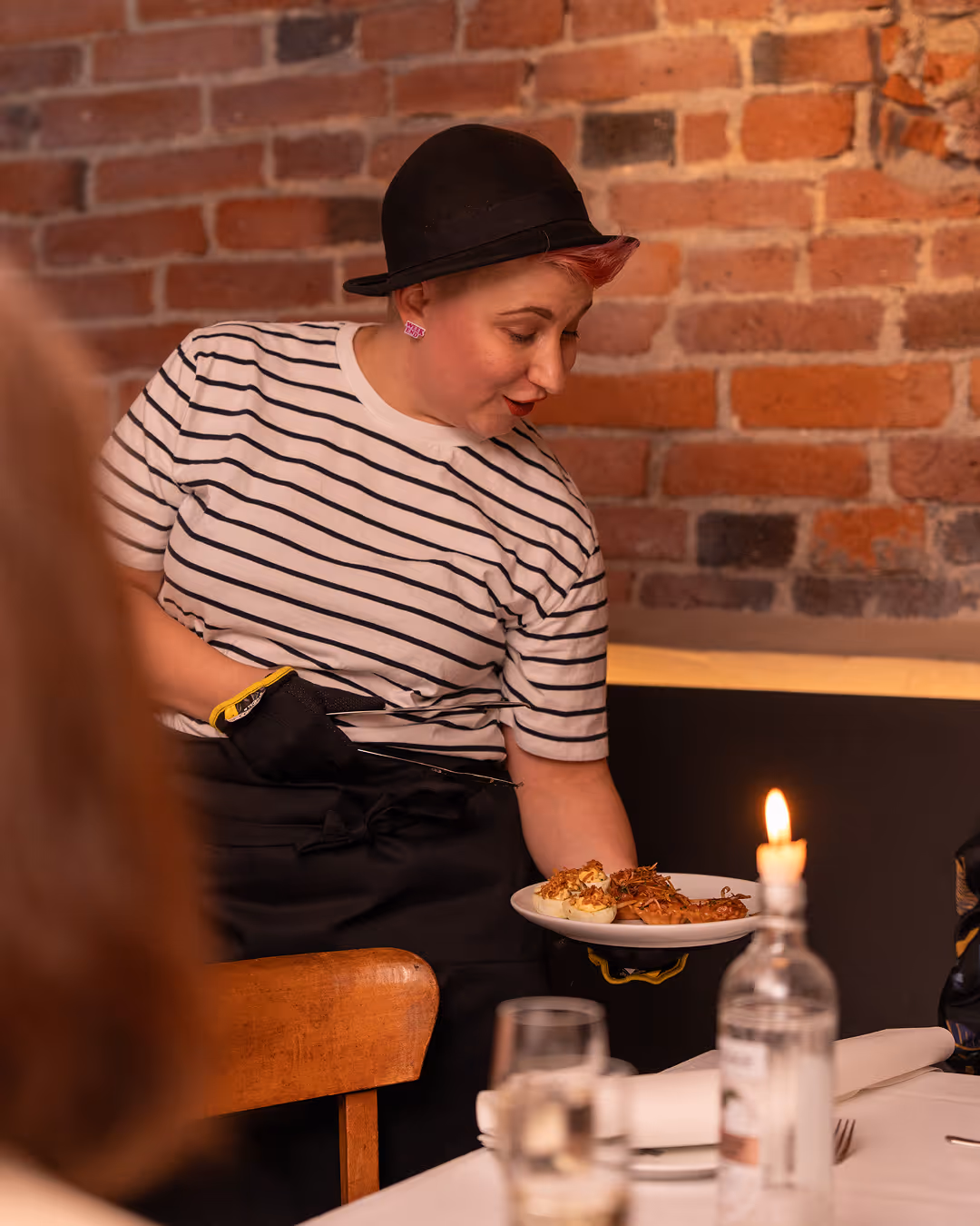 Tarjoilija mustassa hatussa ja raitapaidassa kantaa lautasella ruokia ravintolassa.