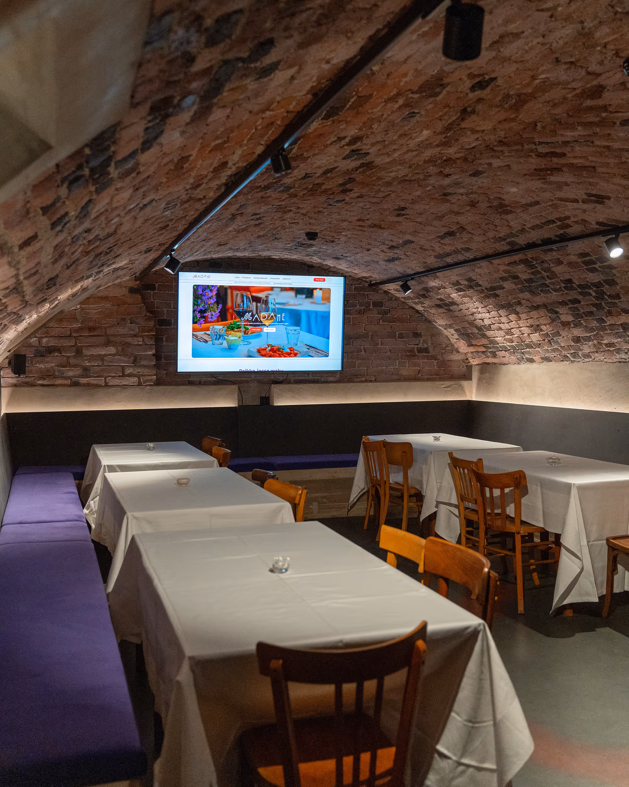 Tunnelimainen huone, jossa on valkoiset liinat peittäviä pöytiä, puisia tuoleja ja seinällä näyttö, joka esittää ravintolasivustoa.