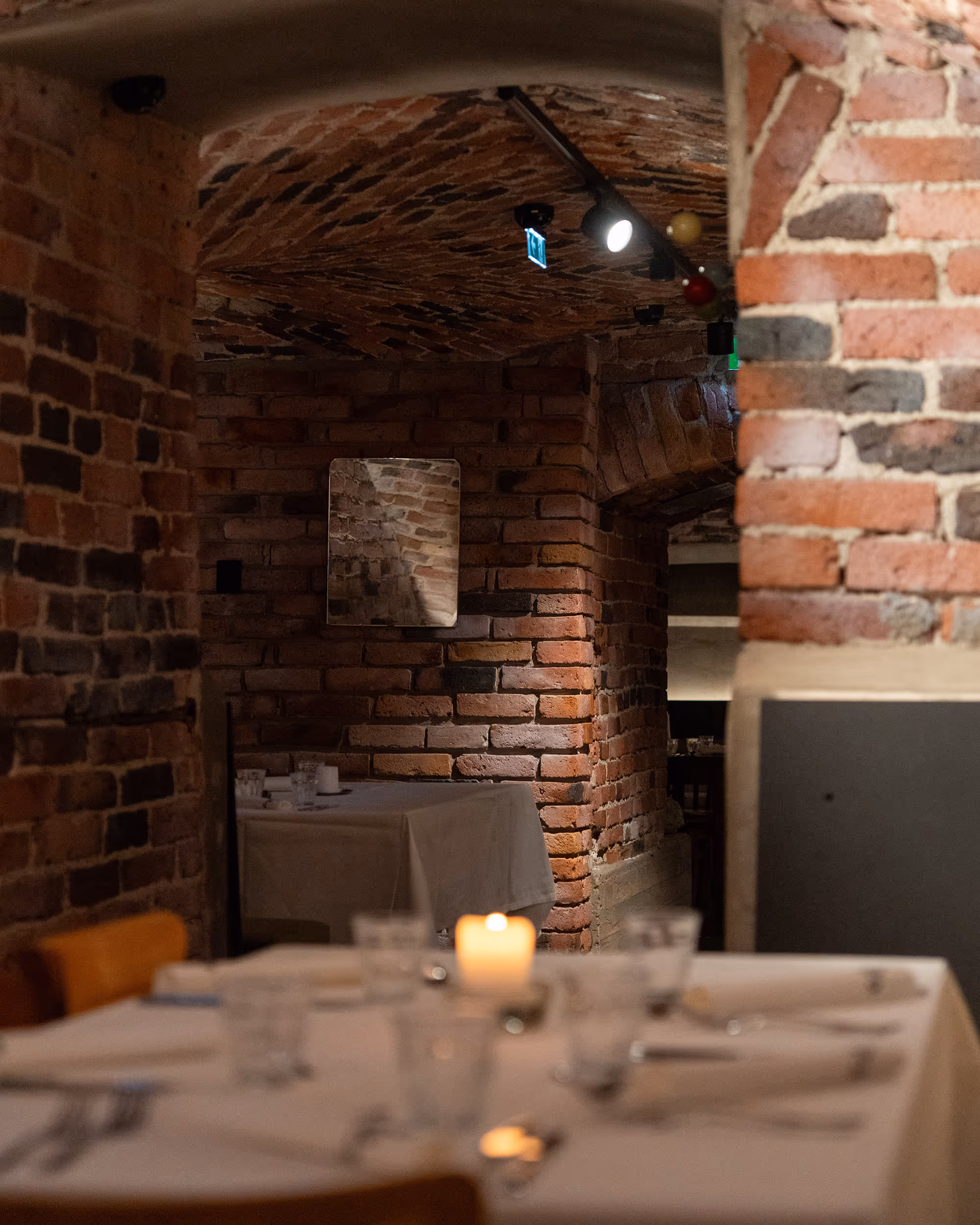 Tunnelinmuotoinen kellariravintolan tila, jossa näkyy tiiliseinät, valkoverhotettuja pöytiä ja kynttilä pöydällä.