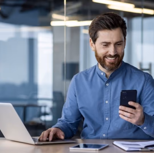 Ein Mann im blauen Hemd, mit einem Handy in der einen Hand und der anderen Hand am Laptop. Er ist ein fröhlicher Mann, der zufrieden mit seiner Payrolllösung ist.