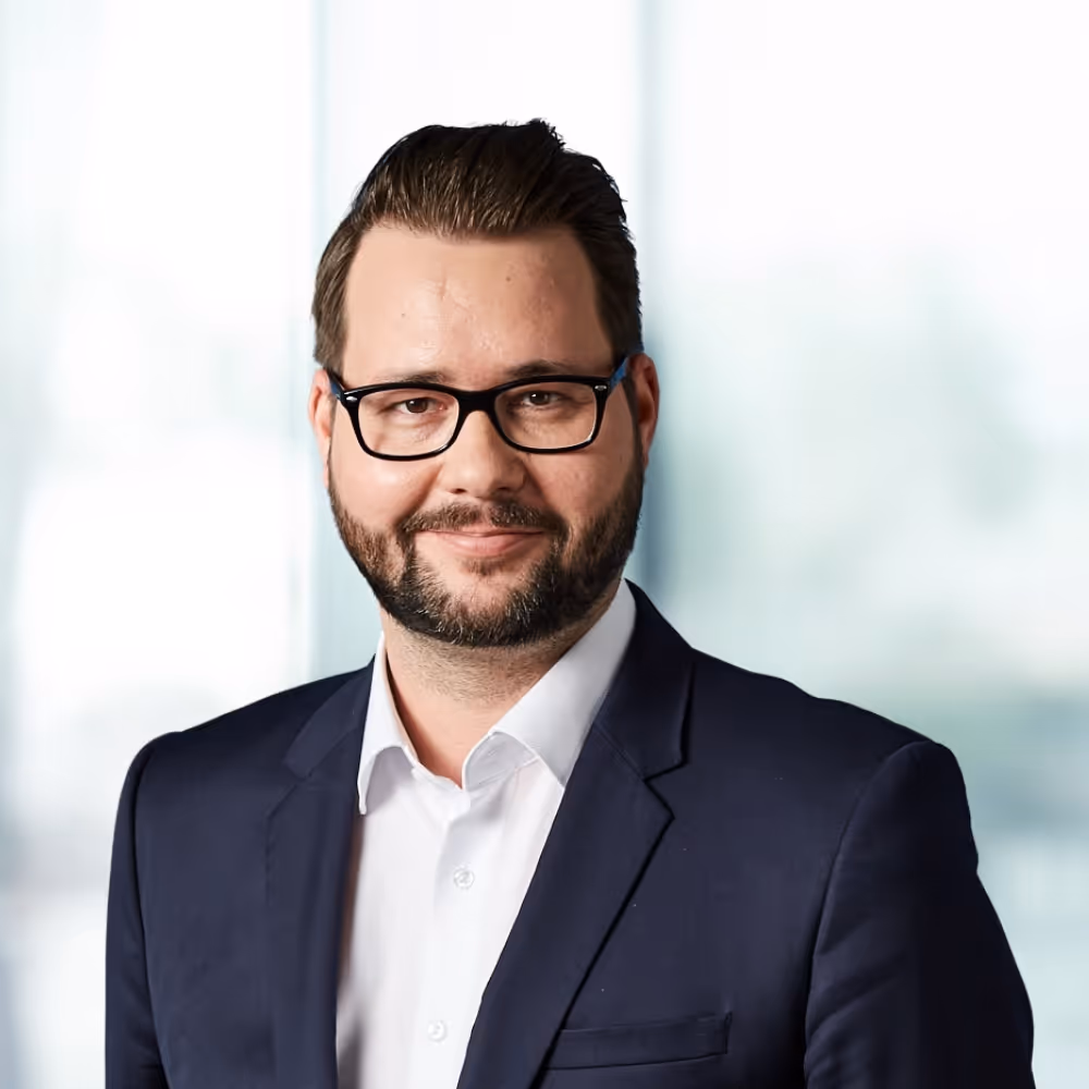 Mann mit Brille, Bart und einem dunklen Anzug vor verschwommenem Hintergrund.