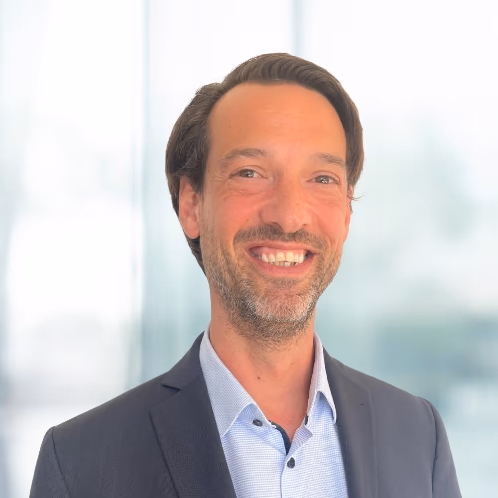 Ein lächelnder Mann mit dunklem Anzug und hellblauem Hemd vor einem verschwommenen Hintergrund.