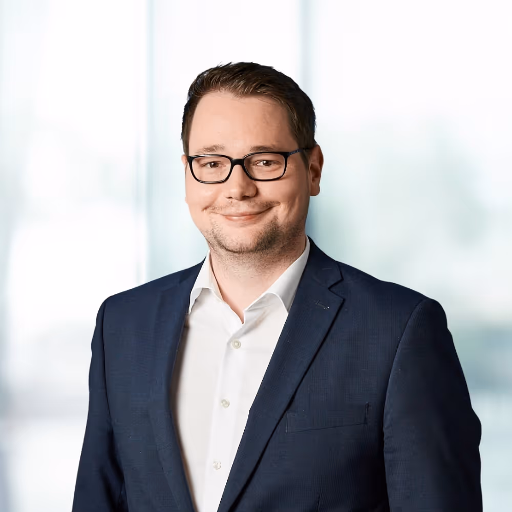 Lächelnder Mann mit schwarzer Brille, weißem Hemd und dunklem Anzug vor unscharfem Hintergrund.