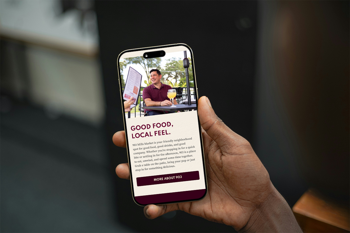 A hand holding an iPhone showing 903 Mills Market's website landing page.