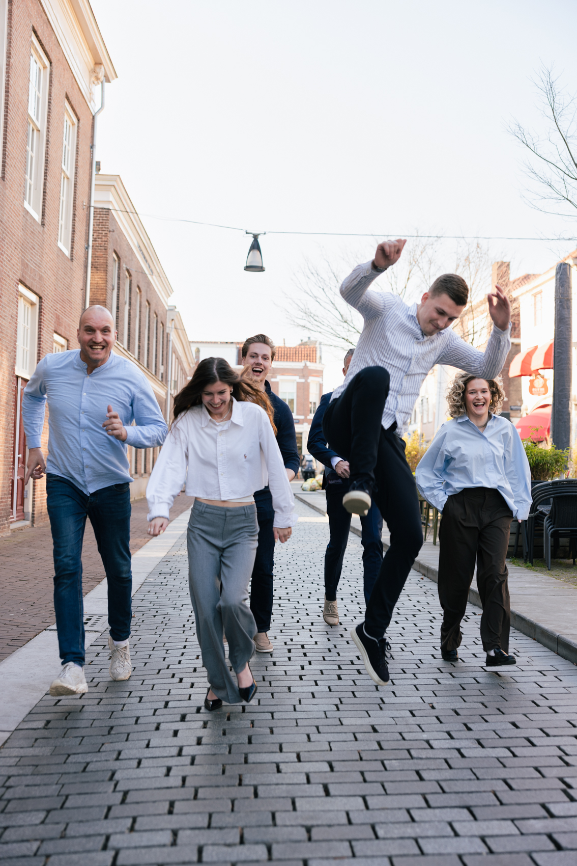 Een groep van zes vrolijke mensen loopt en springt over een met stenen geplaveide straat in een stedelijke omgeving.