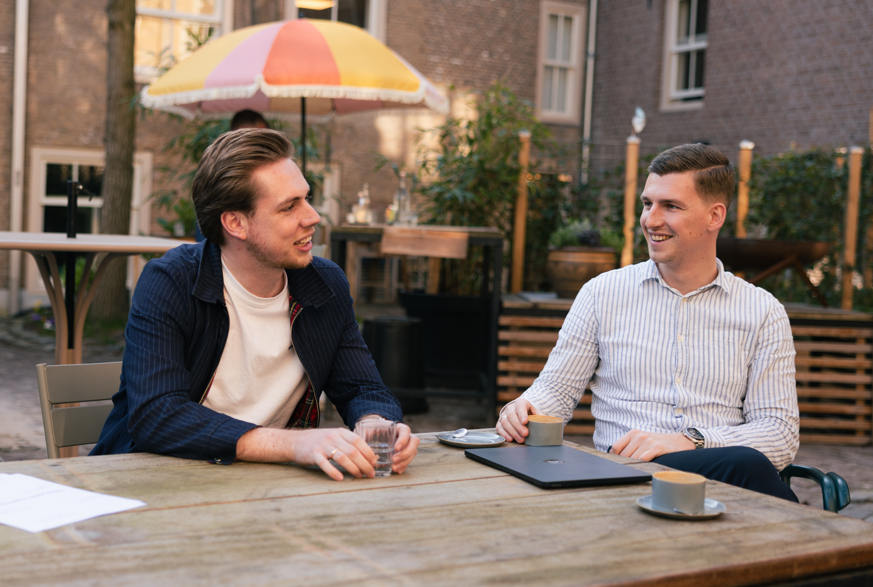Twee mannen zitten buiten aan een houten tafel, pratend en lachend met een laptop en koffiekopjes op de tafel.
