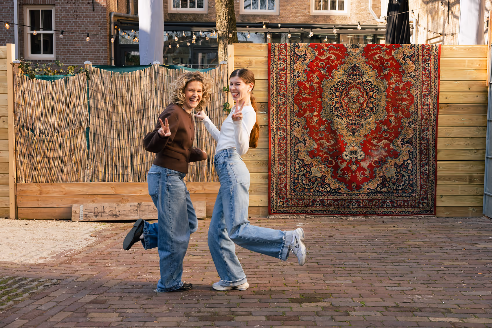 Twee lachende vrouwen die poseren met vredesteken voor een houten schutting en een rode, decoratieve tapijt buiten.