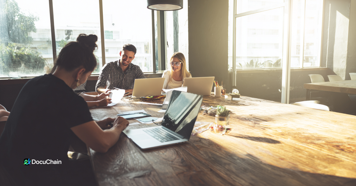 Remote team collaborates around large table with laptops and paperwork, showing friction of signing without digital tools