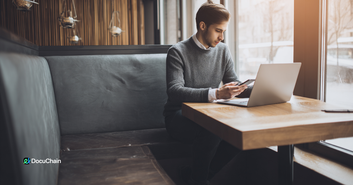 Man uses phone and laptop to review contracts in café booth, illustrating fast and secure signing experience from anywhere.
