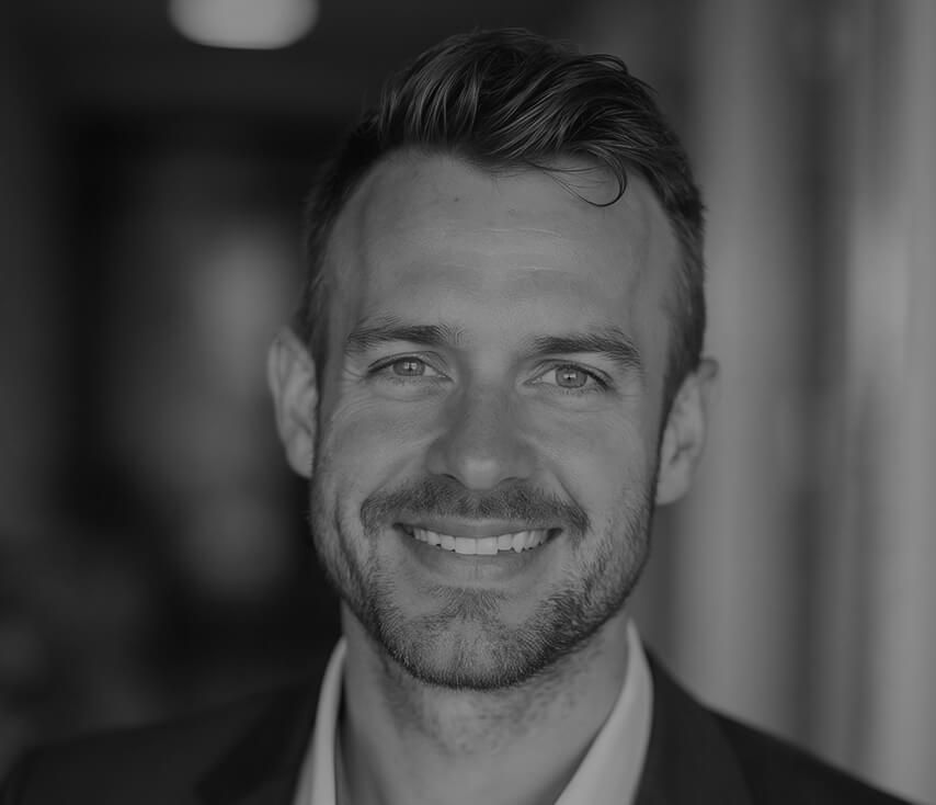 A black and white portrait of a smiling Caucasian man with short hair and a neatly trimmed beard, wearing a suit.