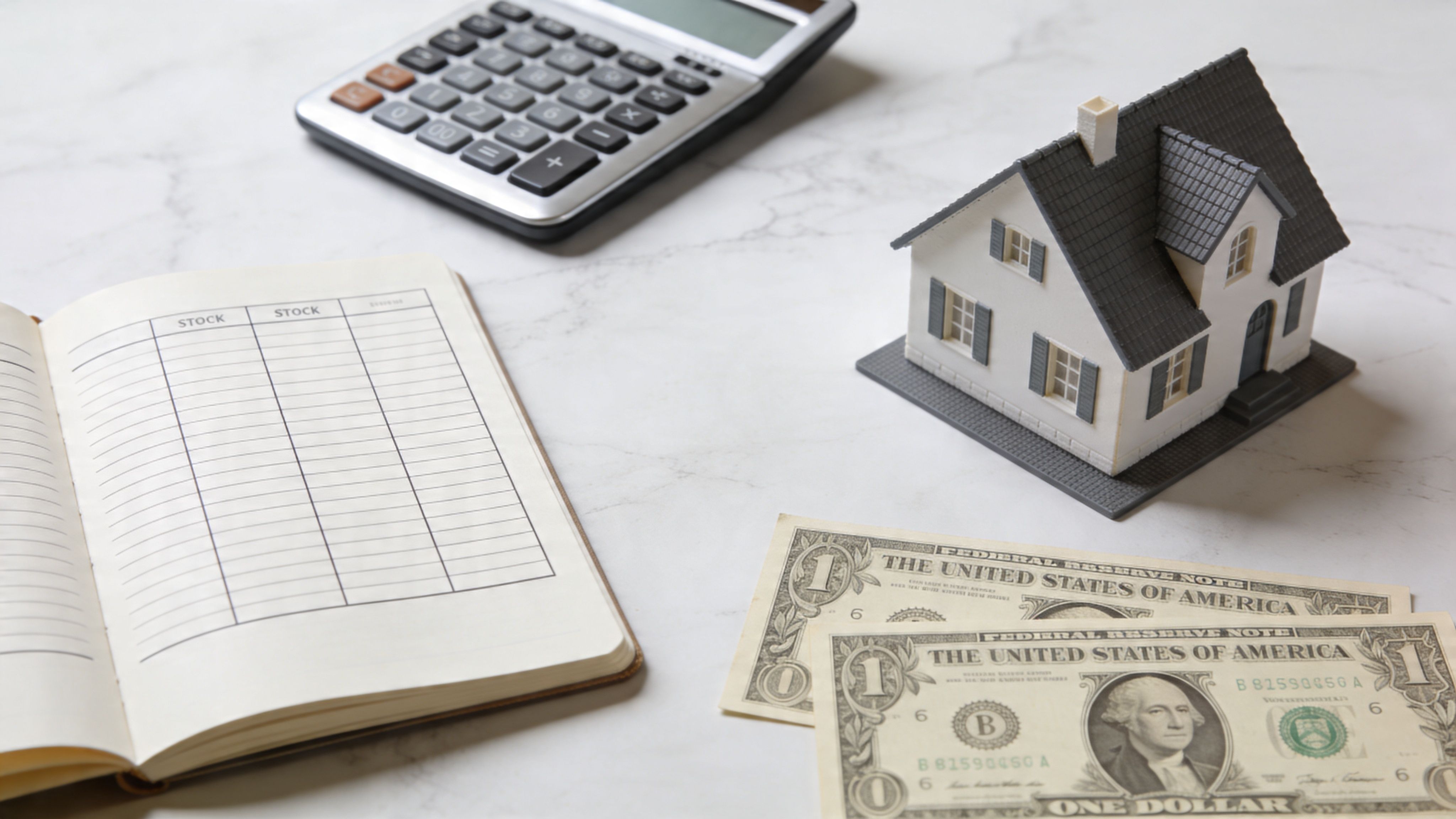 A house model, calculator, open journal with table, and US dollar bills on a marble background.