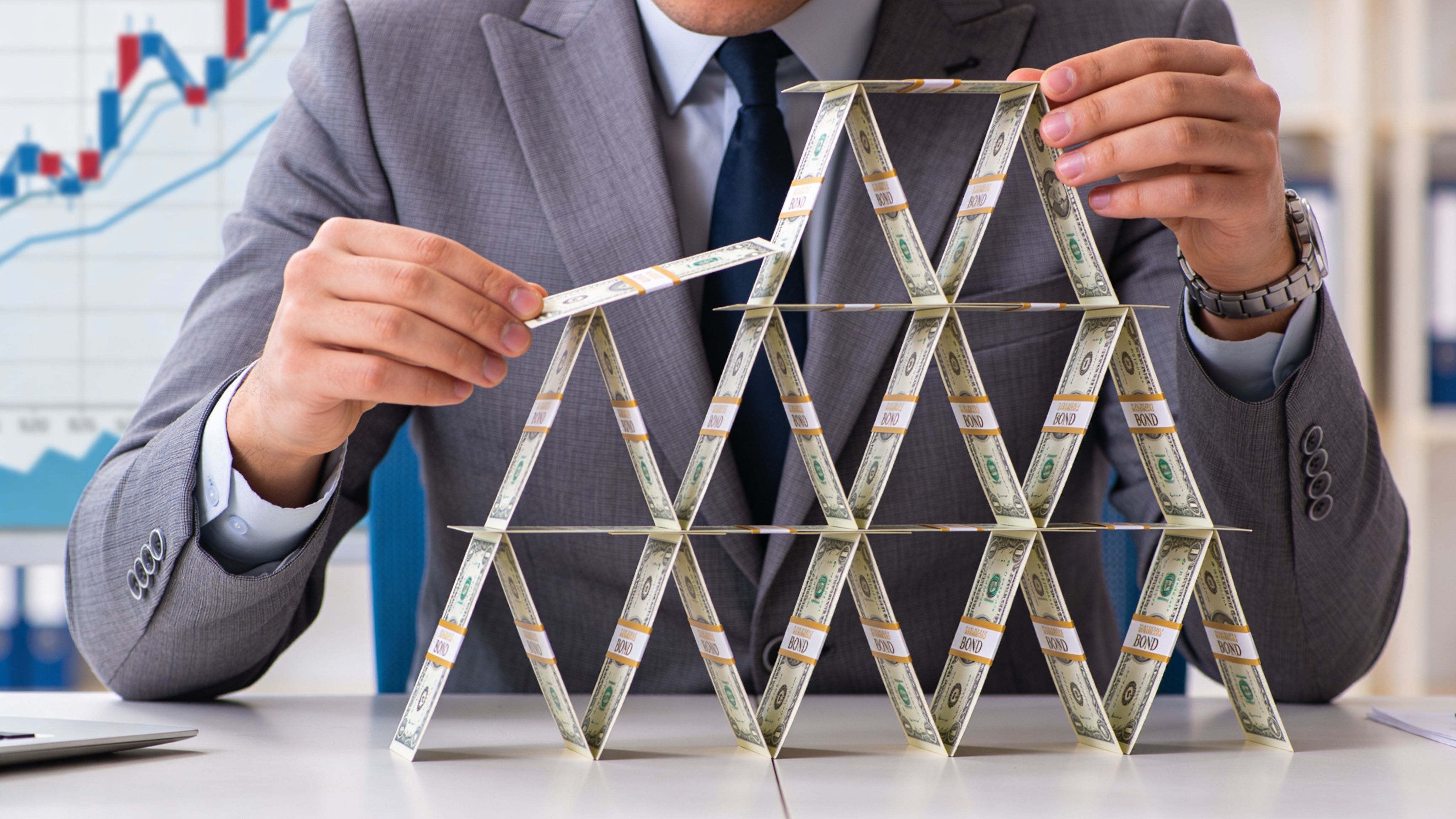 A businessman carefully building a house of cards using stacks of financial bond documents.