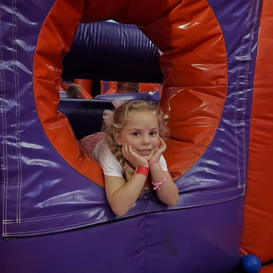 A smiling girl with blonde hair leans on her hands inside a purple and orange inflatable obstacle at an indoor inflatable play park.