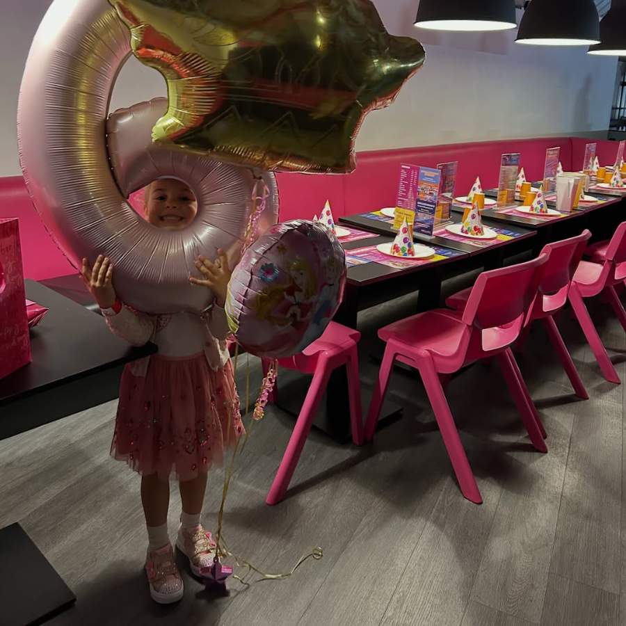 A young girl wearing a pink dress holds large number six balloon in a decorated party area with a long table set up with colourful party hats, menus, and pink chairs.