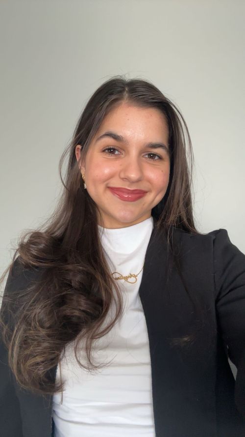 Smiling woman with long dark hair wearing a black blazer and white top against a plain background.