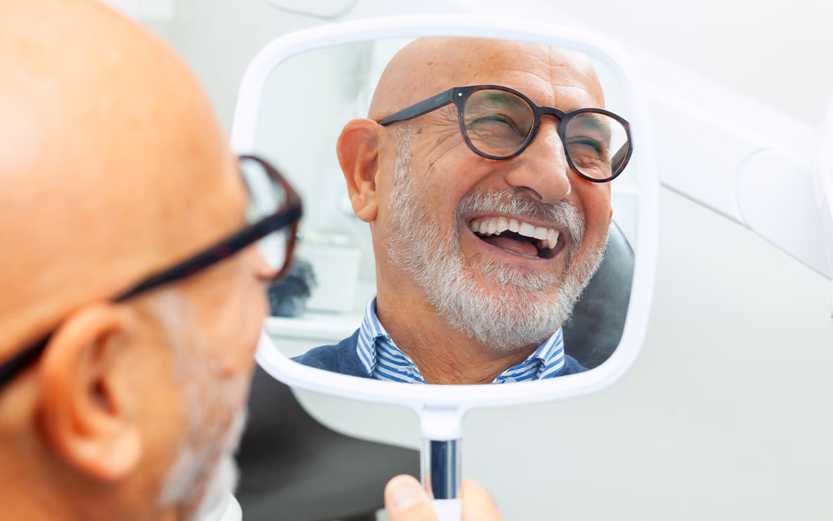 A senior man visiting a top dentist in Los Gatos smiles while looking into a handheld mirror, enjoying his healthy, white smile