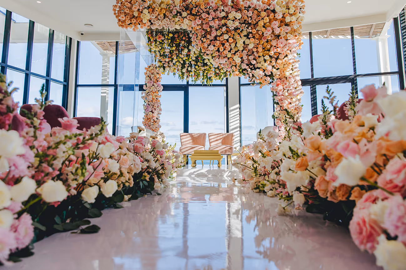 All-inclusive Mexico destination wedding setup with bright wedding aisle lined with pink, white, and peach flowers under a floral arch inside a glass room.