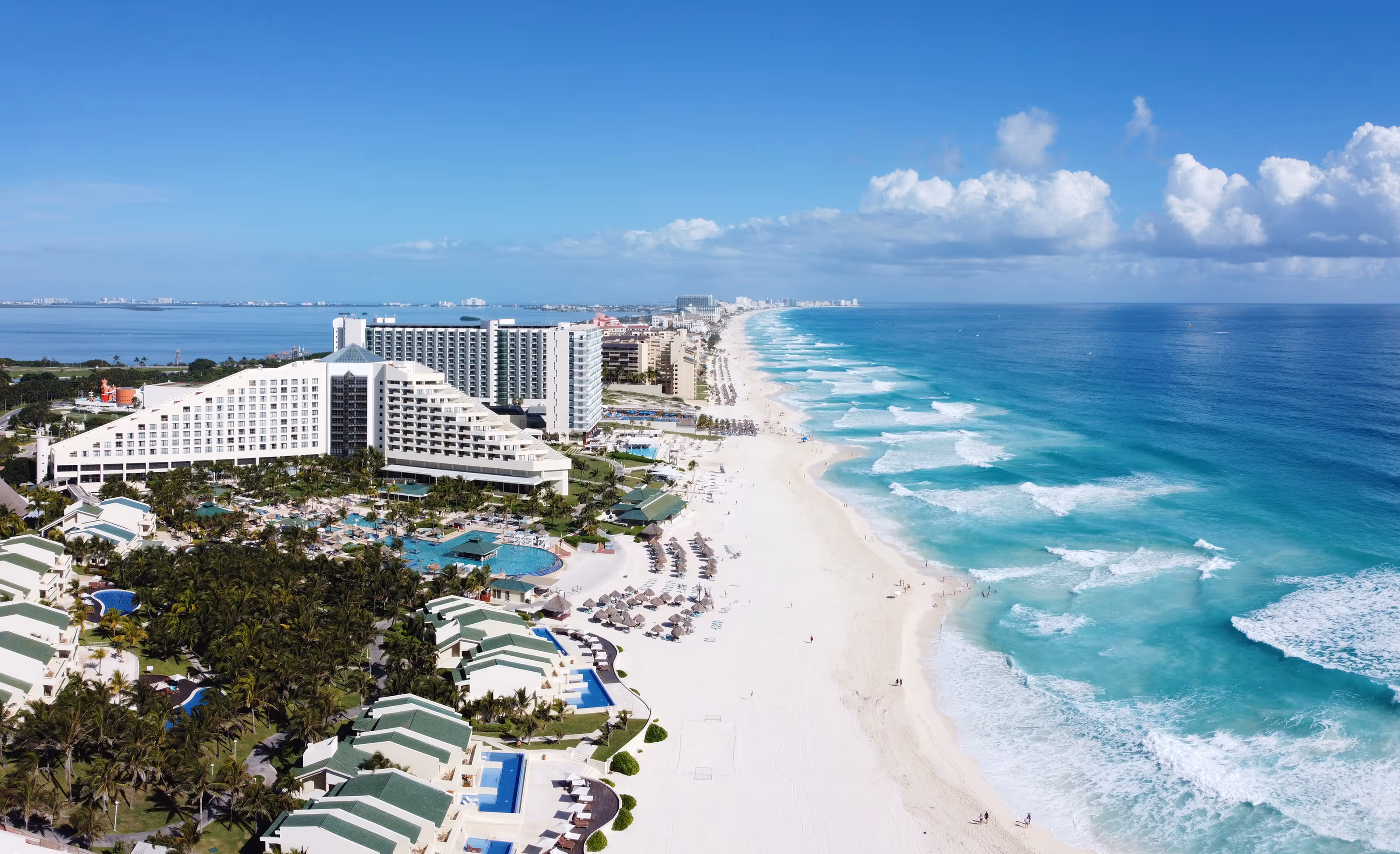 Aerial view of beachfront hotels, white sand beach, and turquoise ocean waves under a cloudy blue sky in Cancun.