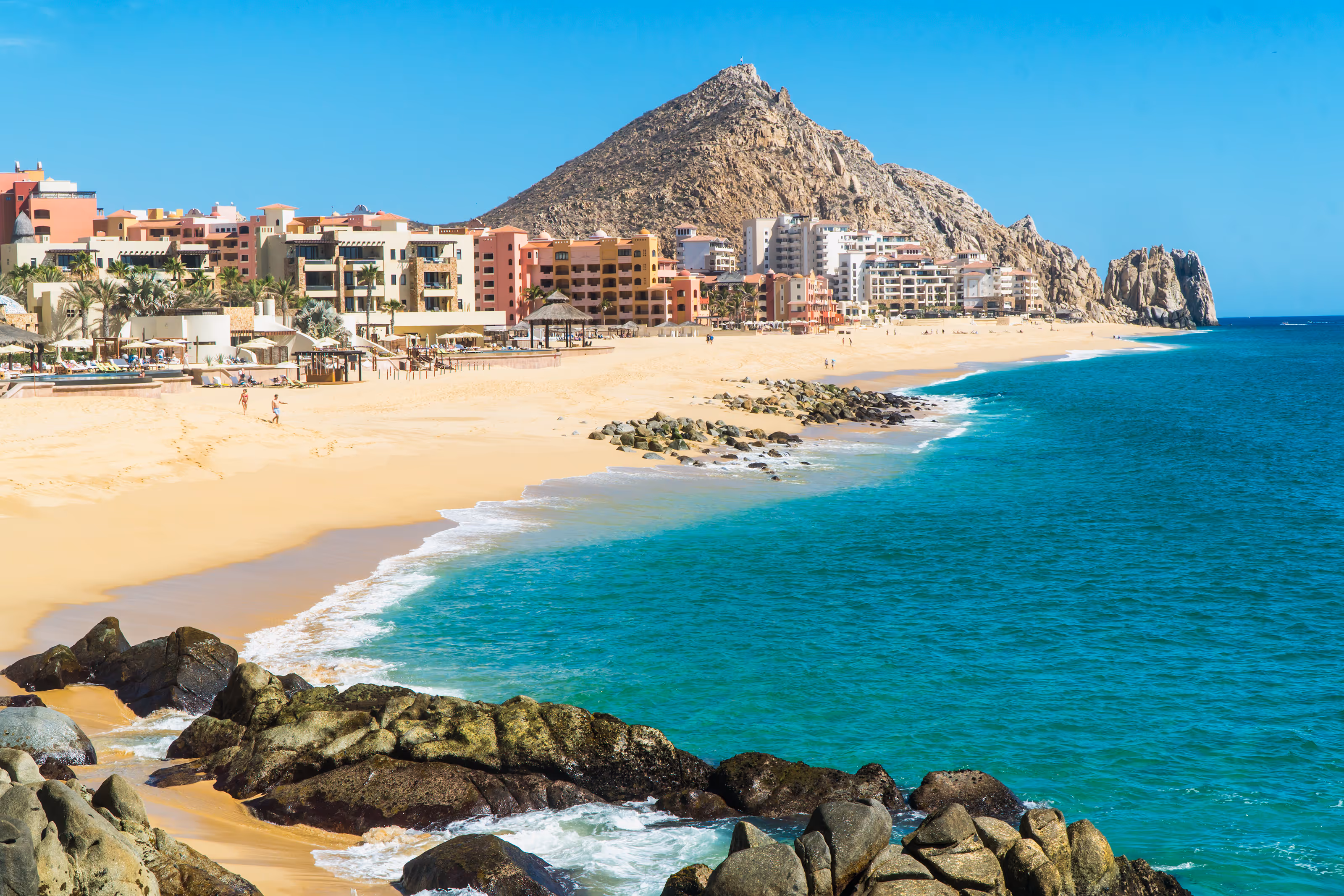 Sunny Los Cabos beach with turquoise water, rocky shoreline, and colourful buildings at the base of a mountain.