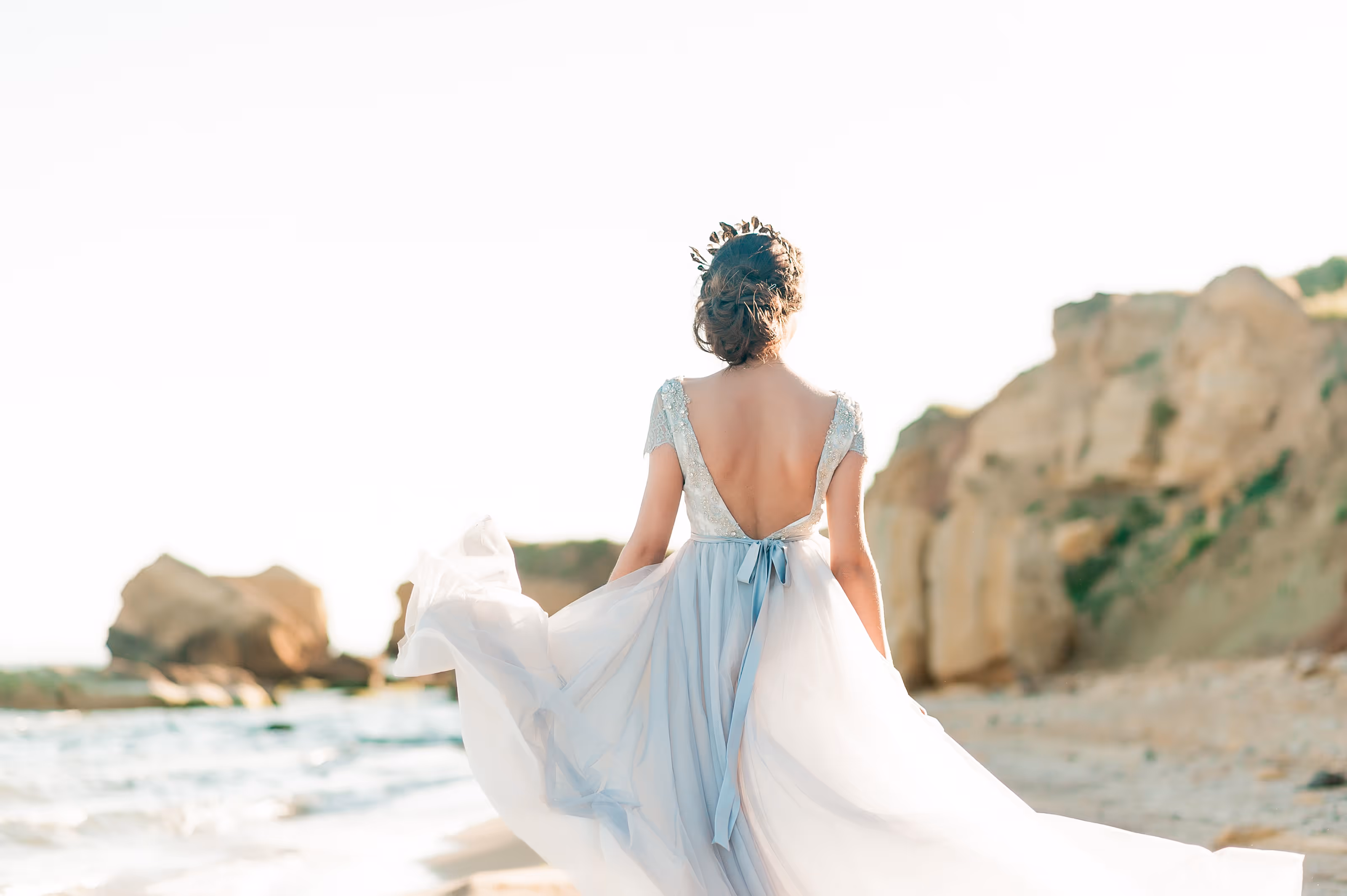Woman in a flowing light blue gown with a low back walking on a beach near rock formations at an all-inclusive Mexico wedding resort.