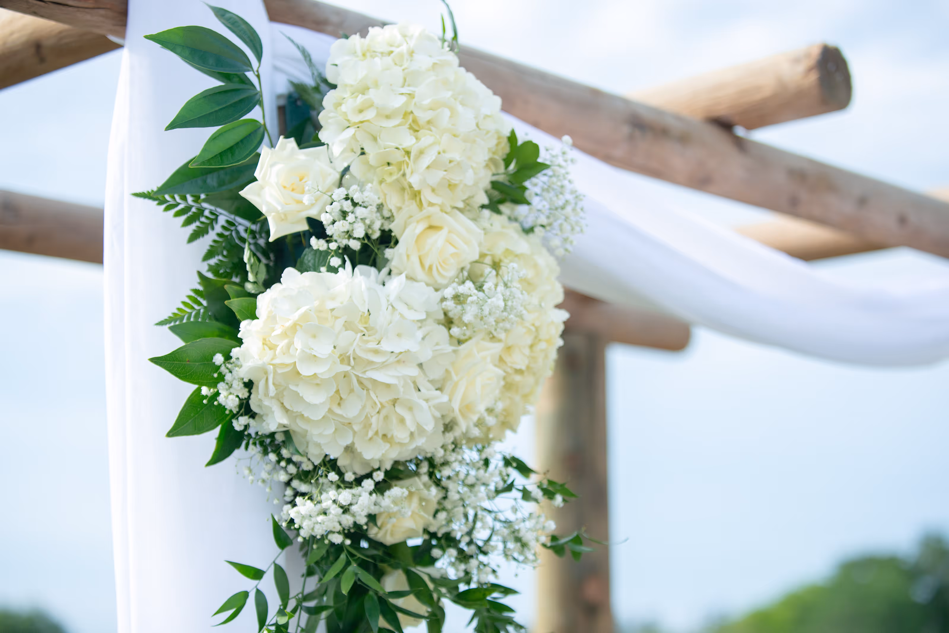 White floral arrangement with roses and hydrangeas on a wooden cross draped in white fabric at a Mexico destination wedding.