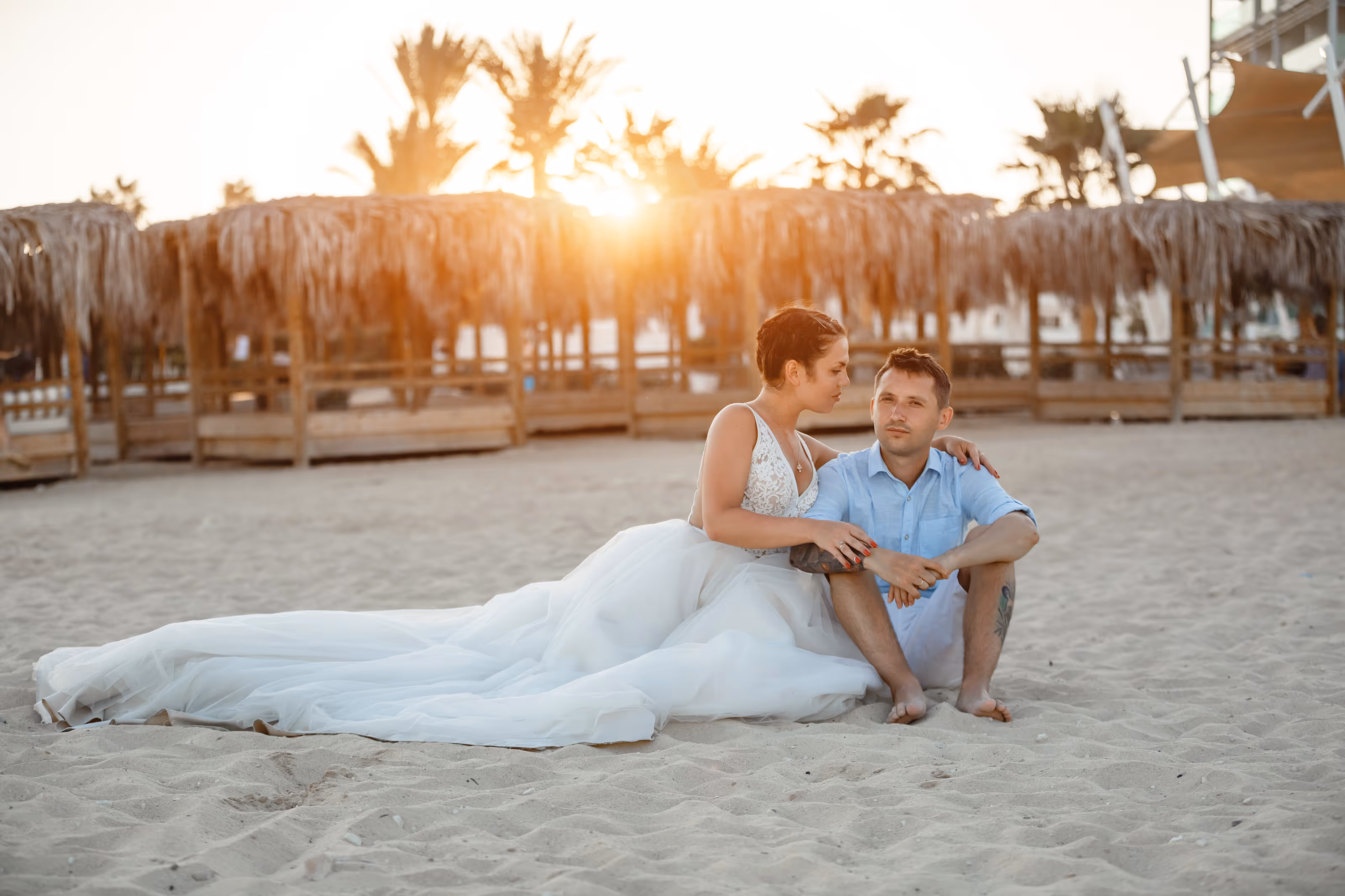 Bride in wedding dress and groom sitting on sandy beach at sunset with thatched umbrellas behind them after their Mexico destination wedding.