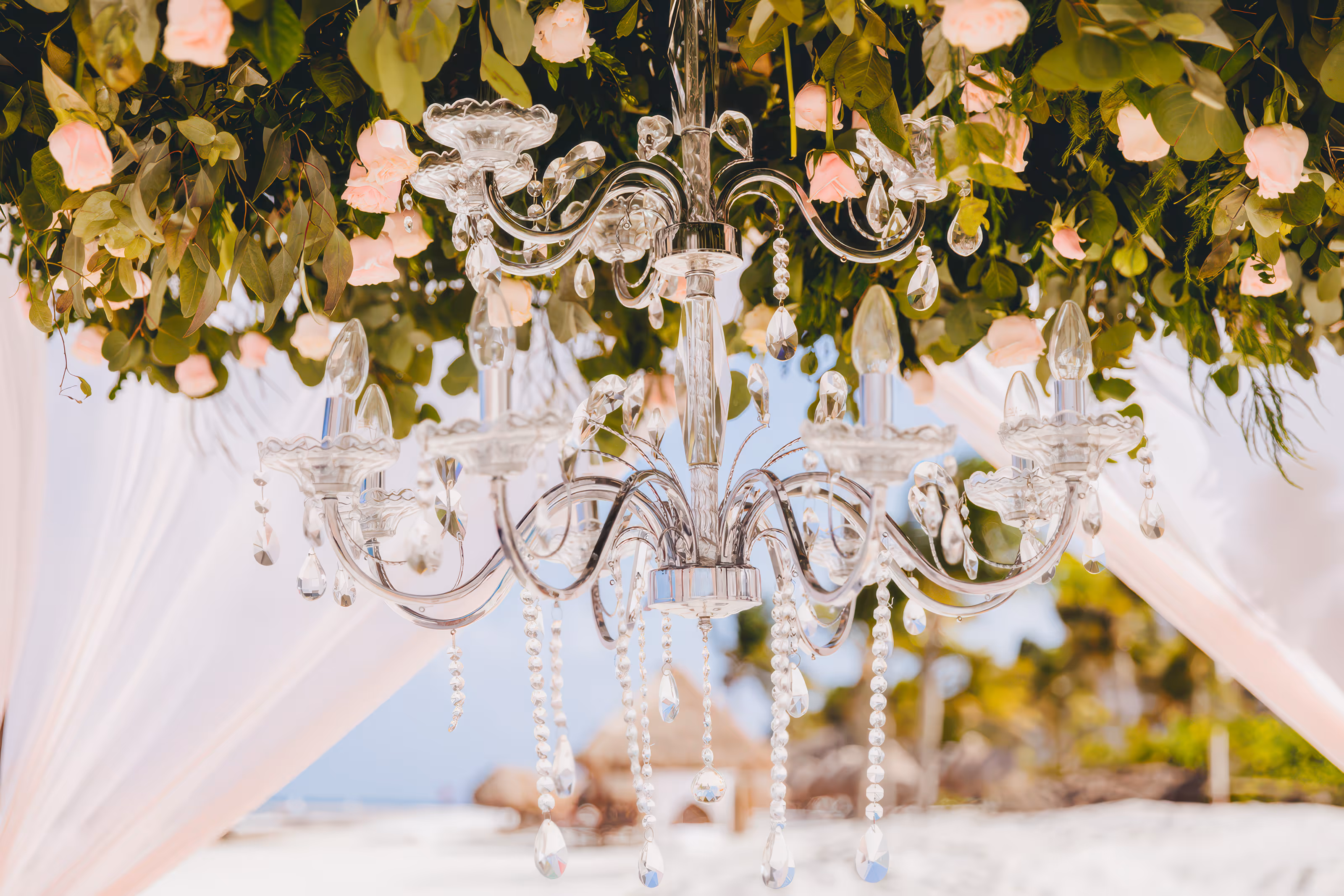 Crystal chandelier hanging beneath green leaves and pink rose flowers in an outdoor setting during a Mexico destination wedding.