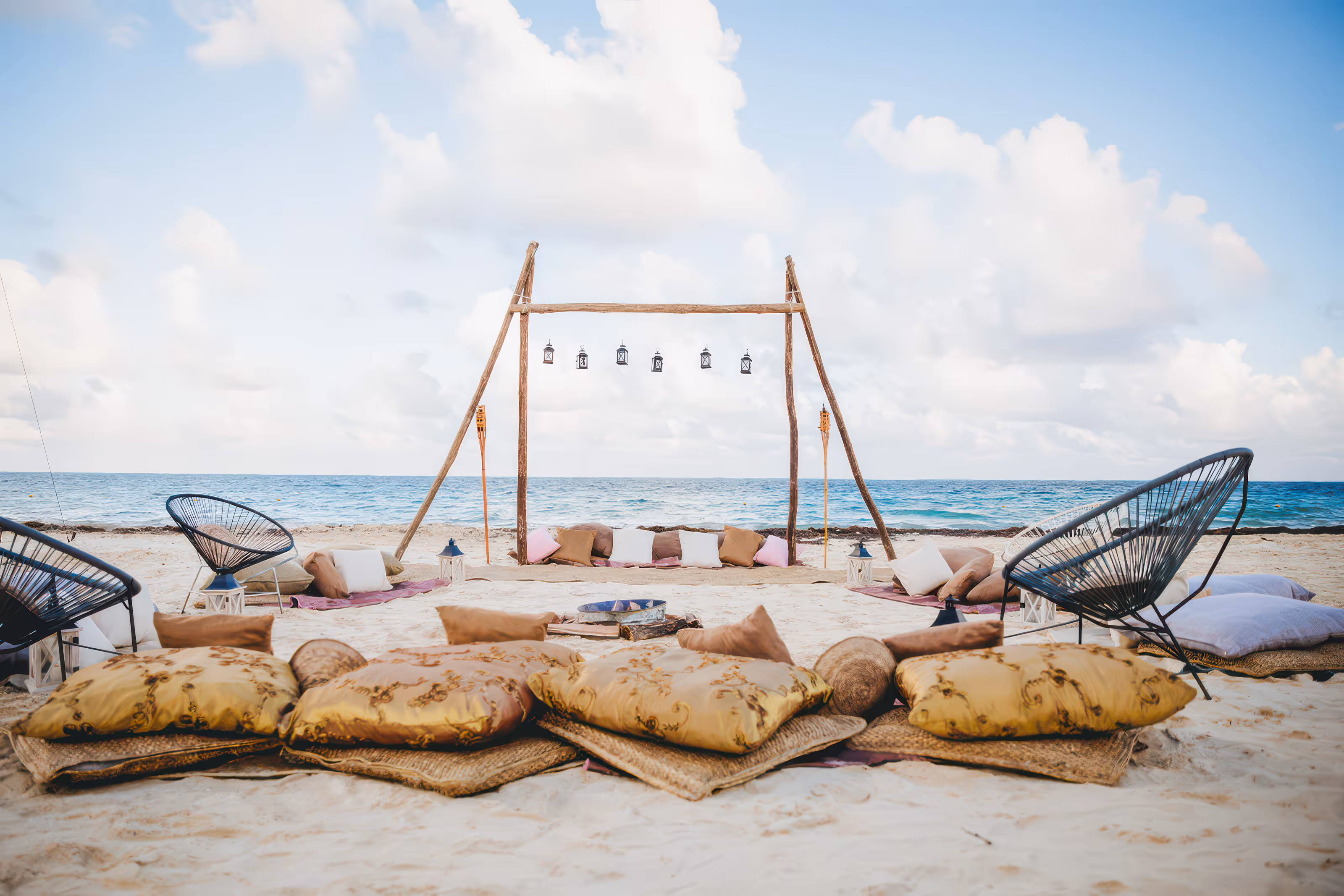 Mexico destination wedding beach seating setup with cushions, chairs, lanterns, and a wooden frame by the ocean on sand.