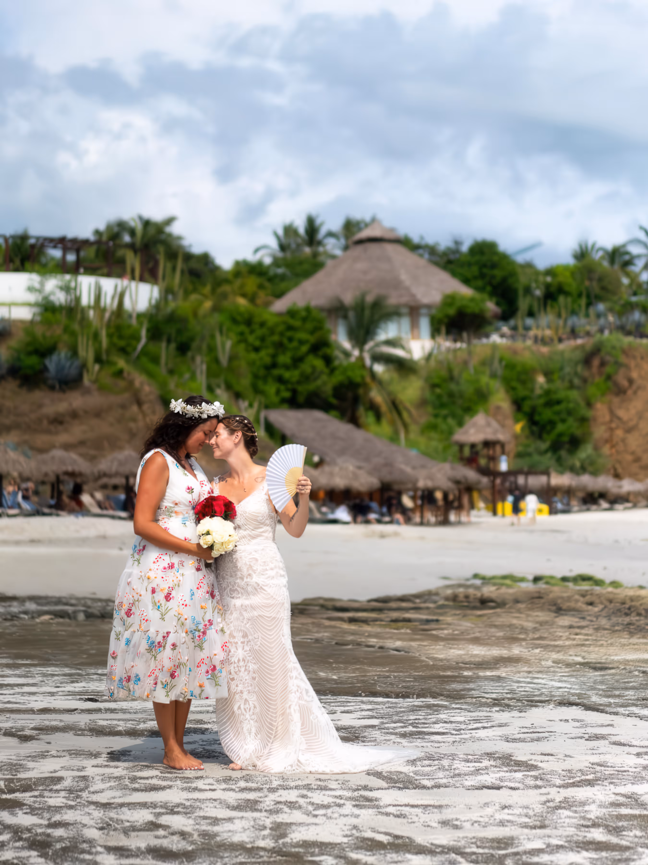 Two brides in wedding dresses stand barefoot close on a beach holding bouquets and a fan after their Mexico destination wedding.