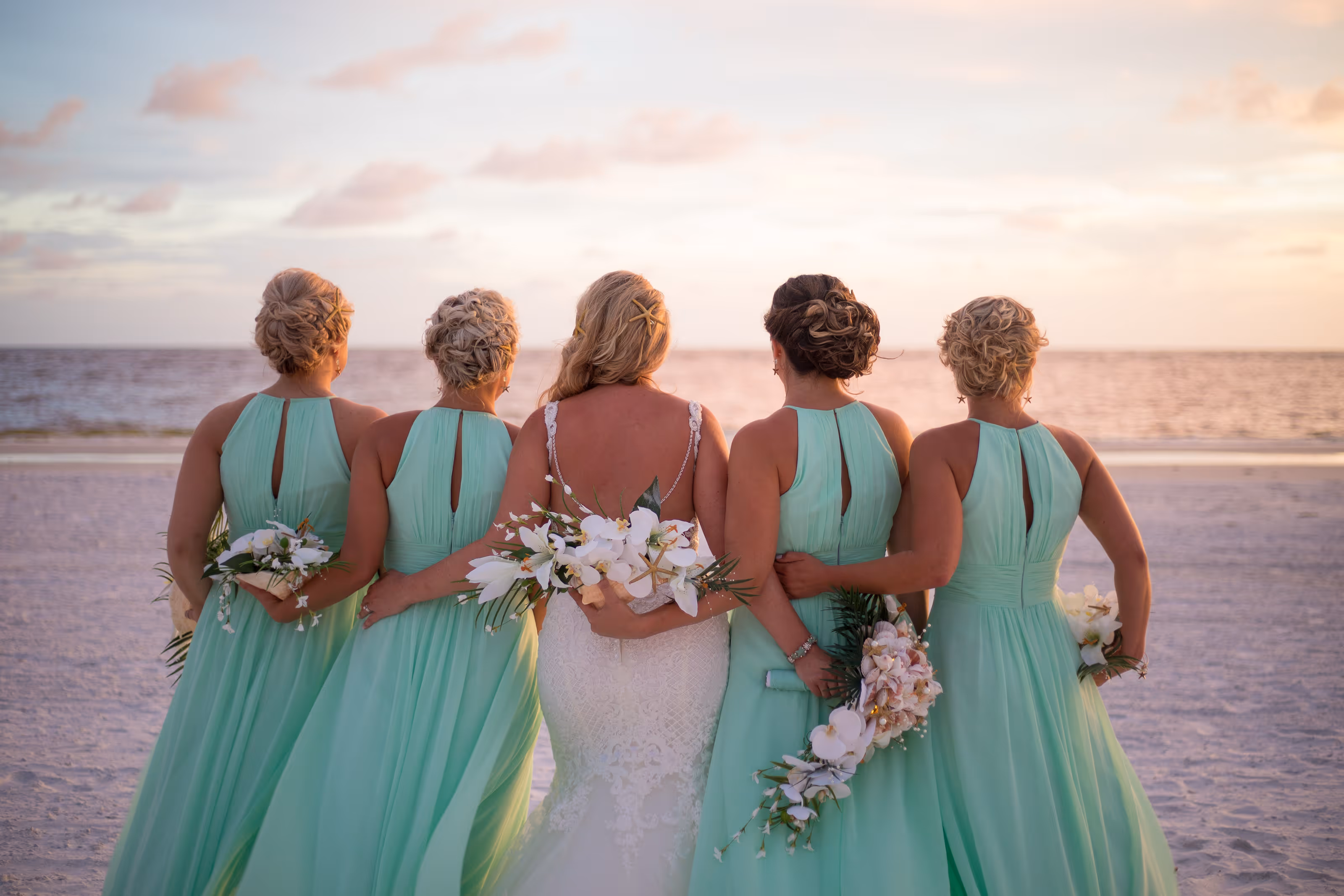 Bride in white dress and four bridesmaids in mint dresses standing arm in arm on a beach at sunset after a Dominican Republic destination wedding.