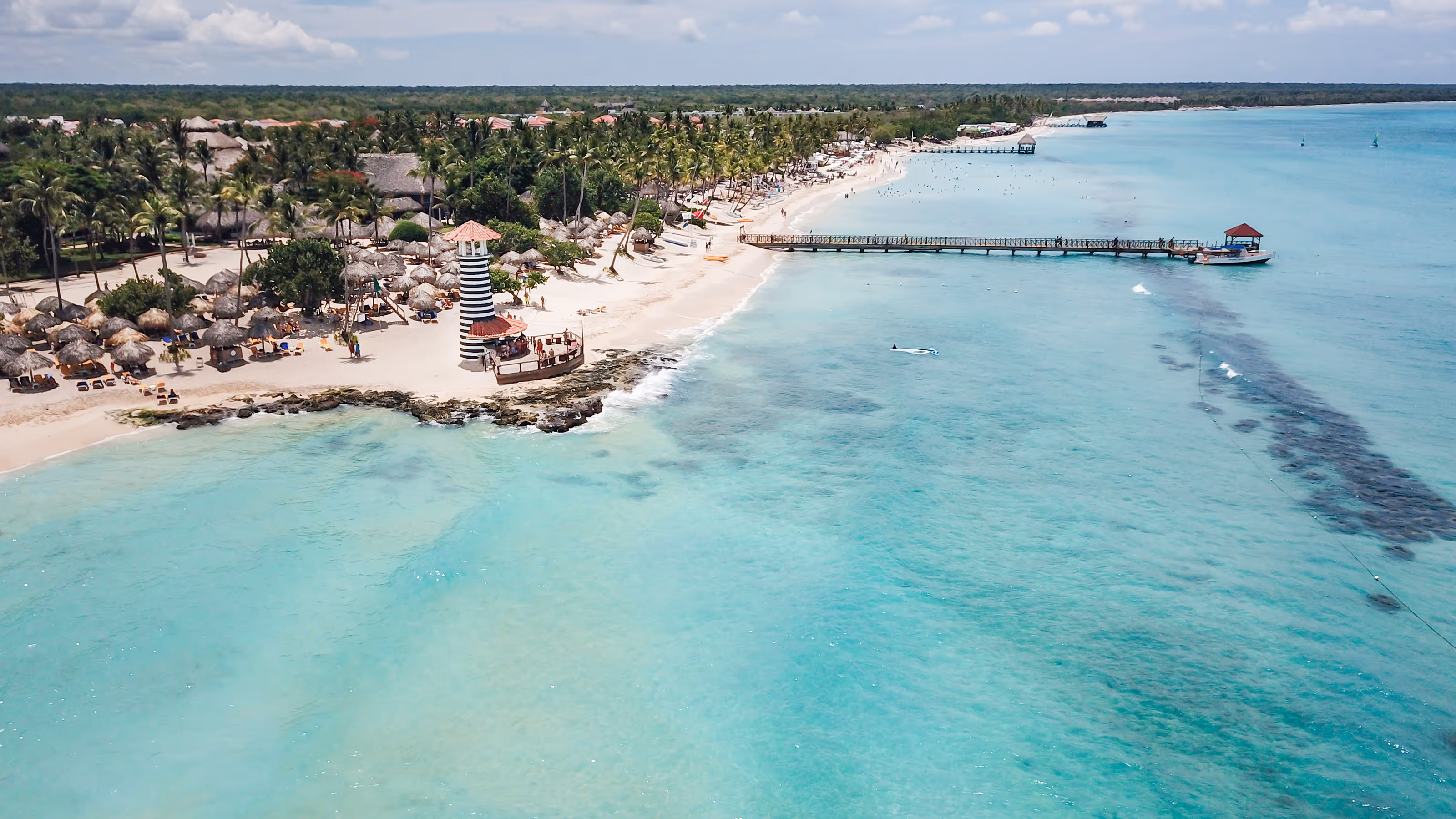 Aerial view of an all-inclusive Dominican Republic wedding resort with palm trees, a striped lighthouse, piers, and turquoise water.
