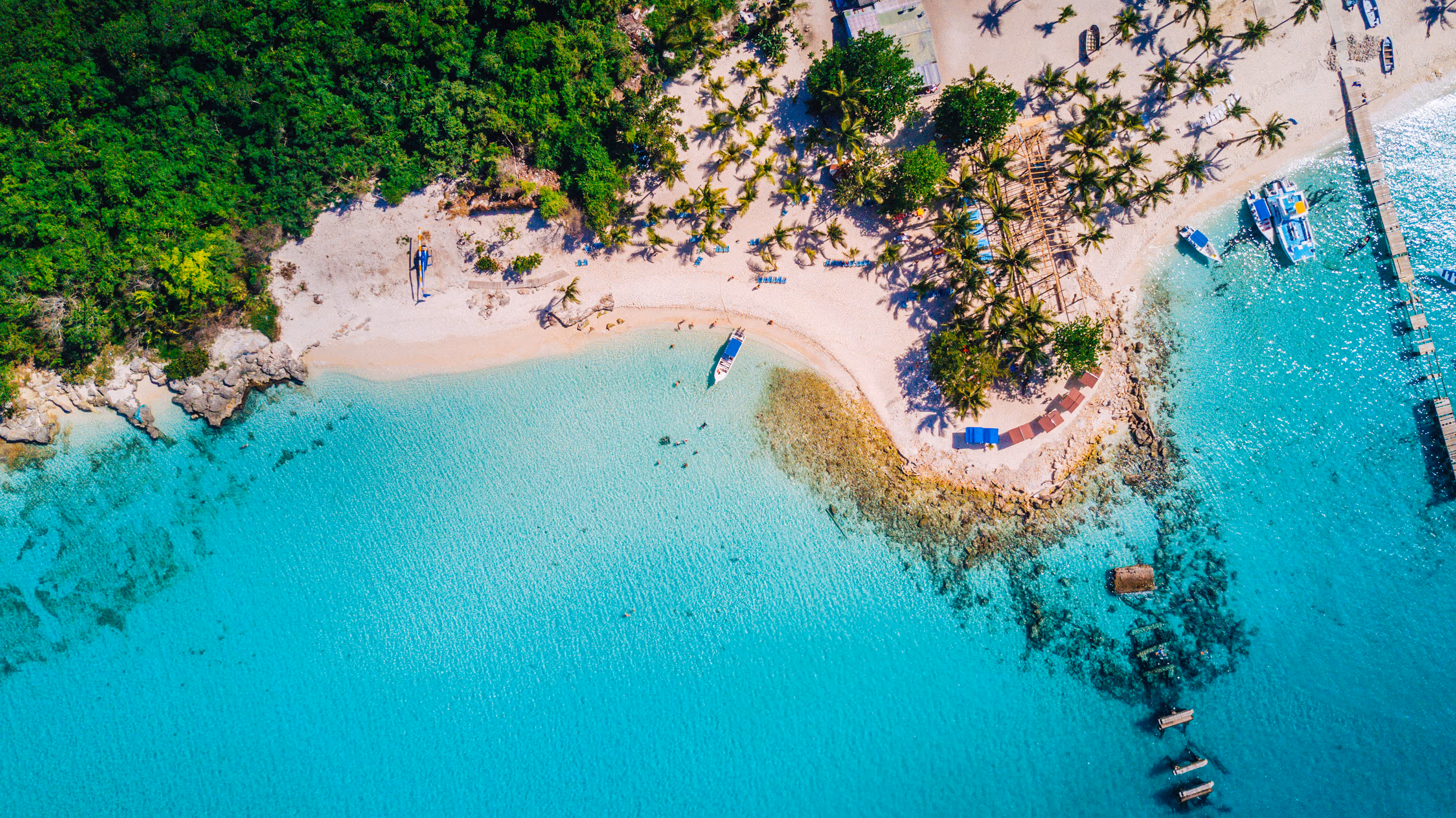 Aerial view of turquoise water, sandy beach, palm trees, boats, and forest on the coast of Punta Cana at a destination wedding resort.
