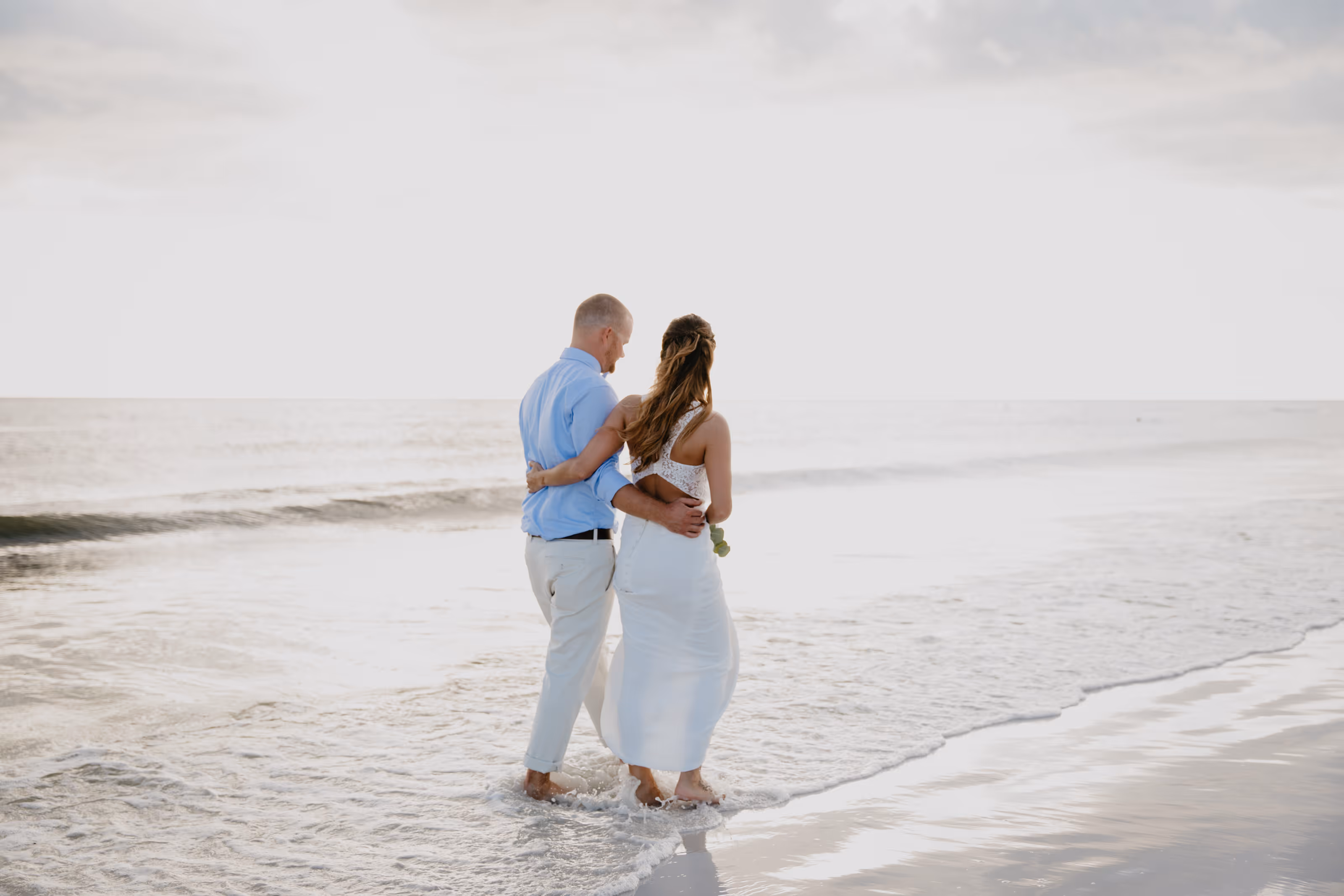 Couple in white and light blue attire walking barefoot at the beach with ocean waves after their Dominican Republic destination wedding.