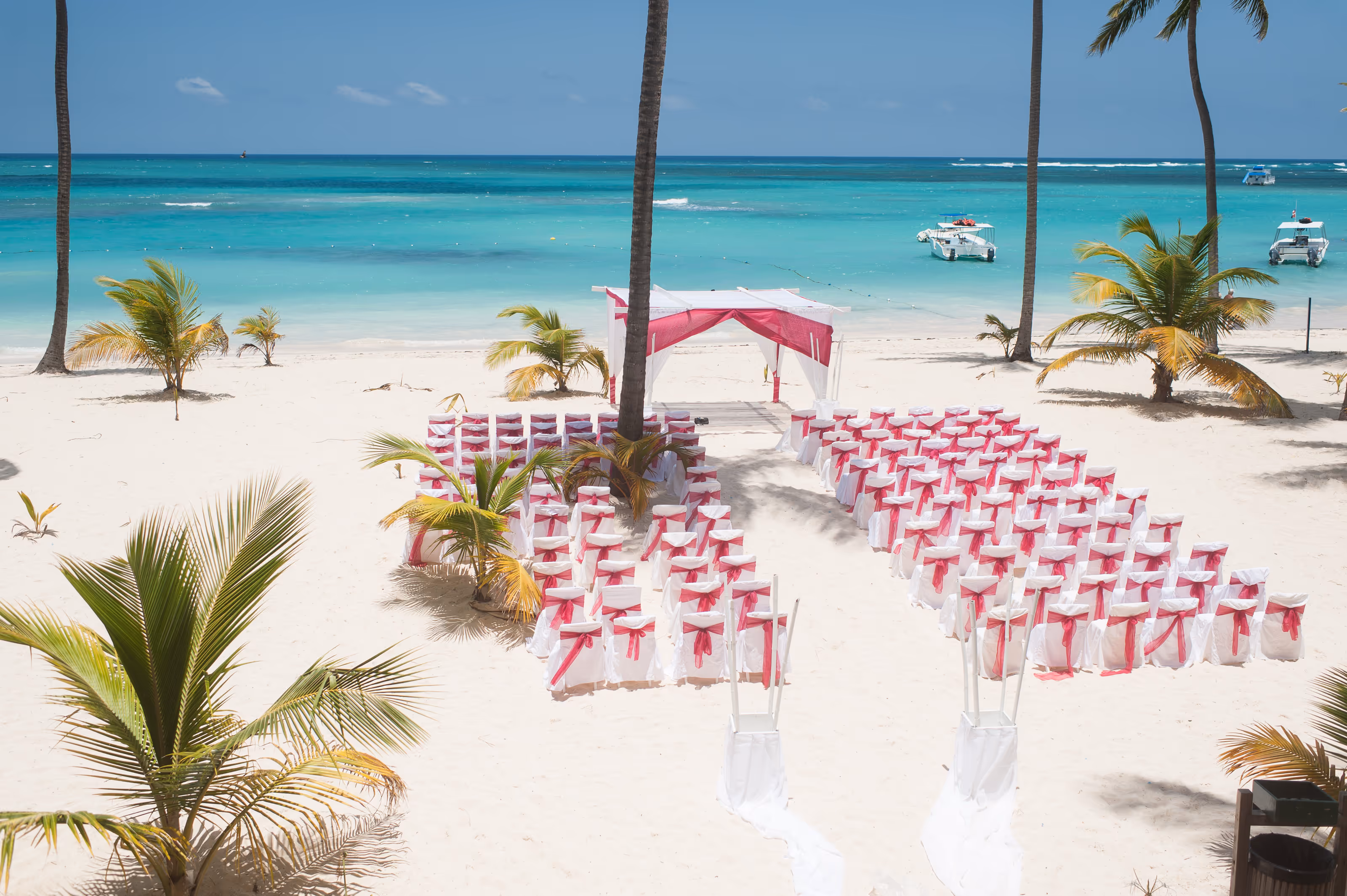 Dominican Republic destination wedding setup with white chairs and pink bows facing an altar on the beach near turquoise ocean and palm trees.