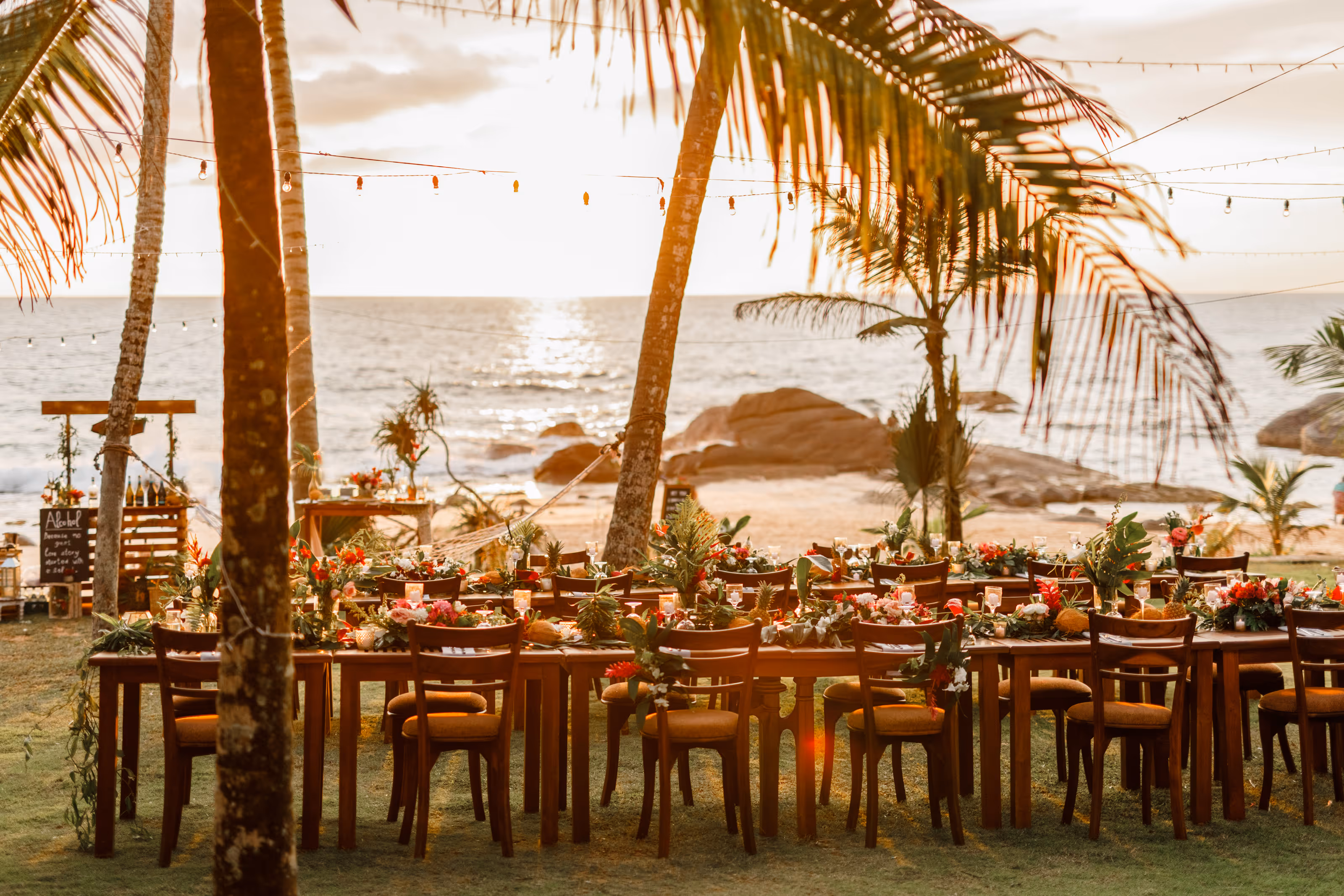 Sunset beach dining setup with wooden tables and chairs decorated with flowers and pineapple centerpieces at a Dominican Republic destination wedding.