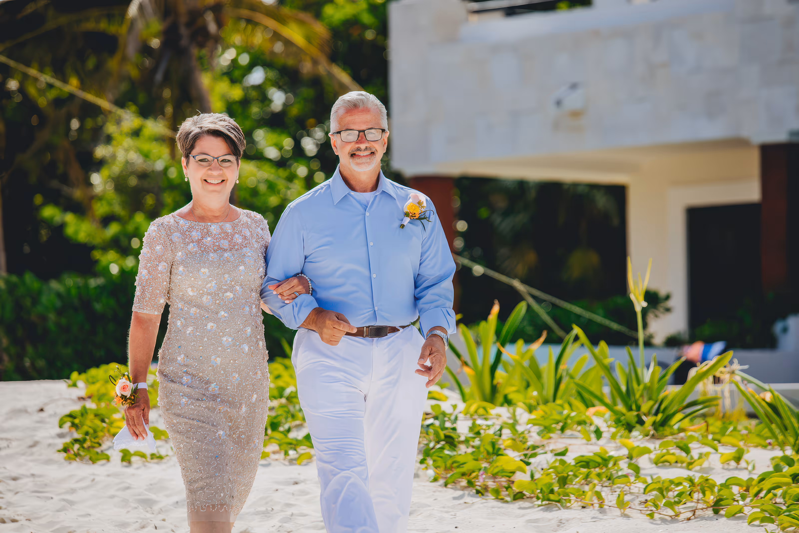Smiling guests of a Dominican Republic destination wedding walking arm in arm on sandy path with greenery and building in background.