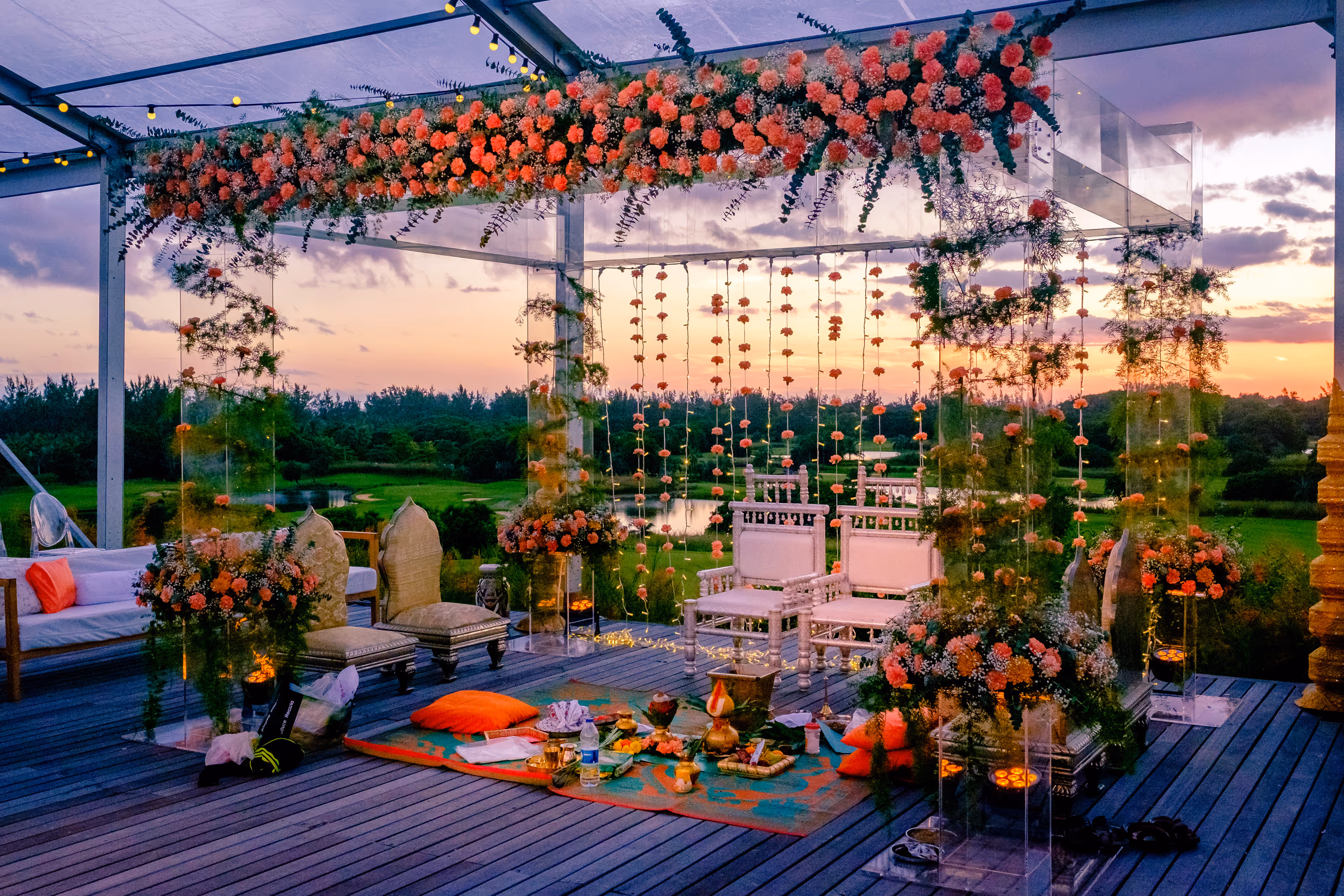 Outdoor Dominican Republic destination wedding mandap decorated with orange flowers, white chairs, and cushions at sunset.