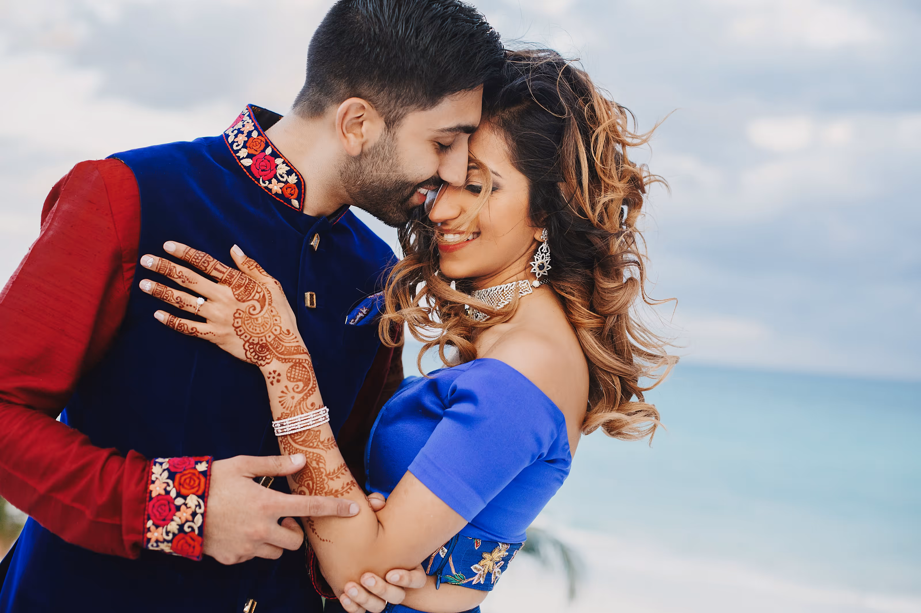 South Asian couple embracing with smiles, woman in blue dress and mehndi on her hand near a beach at an all-inclusive destination wedding resort.