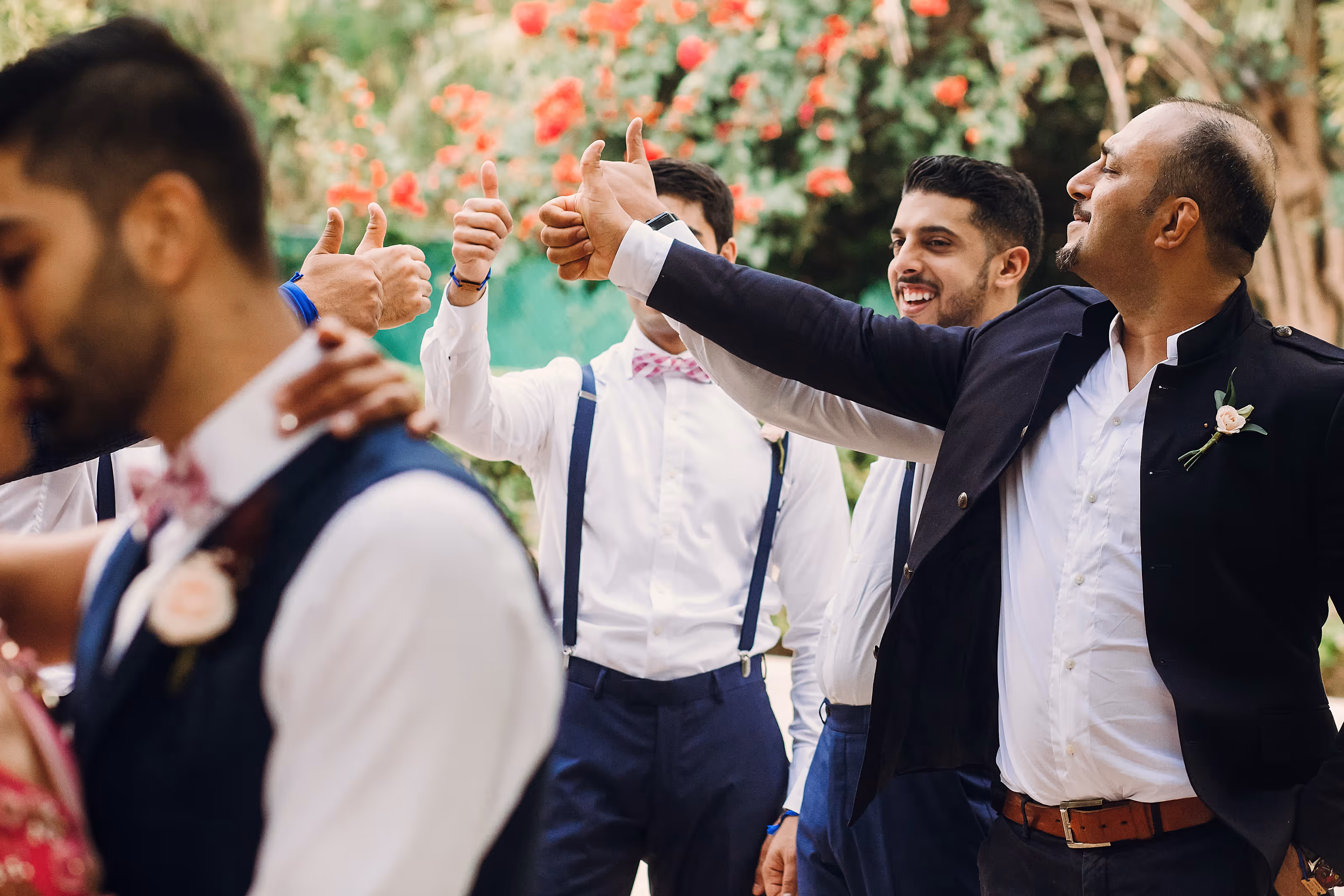 Group of men in formal attire smiling and giving thumbs up outdoors with red flowers in background at a South Asian destination wedding.