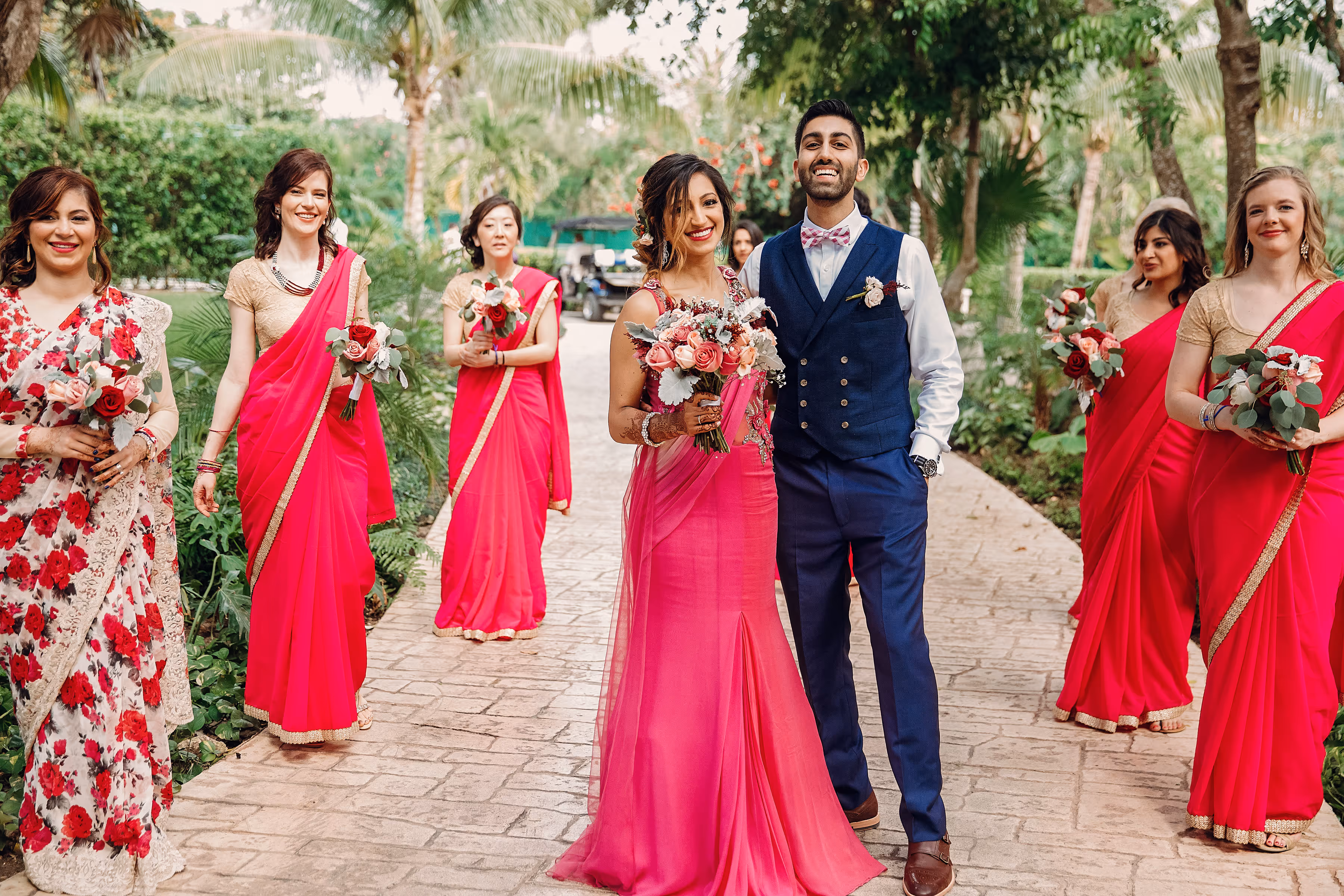 Bride in pink gown and groom in blue suit with bridesmaids wearing bright pink saris outdoors at a South Asian destination wedding.