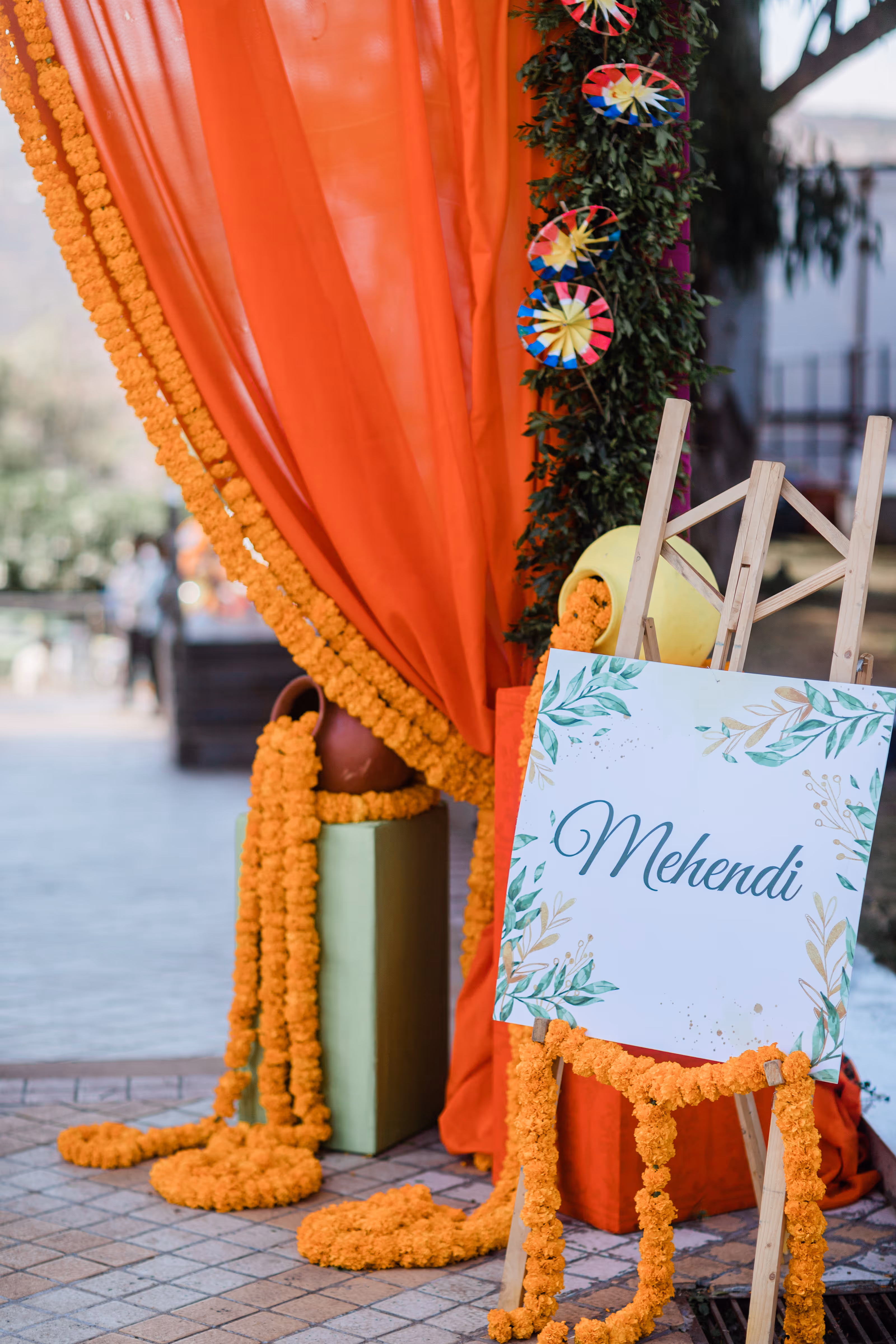 Decorated mehendi destination wedding sign with marigold garlands and orange drapes on an easel outside.