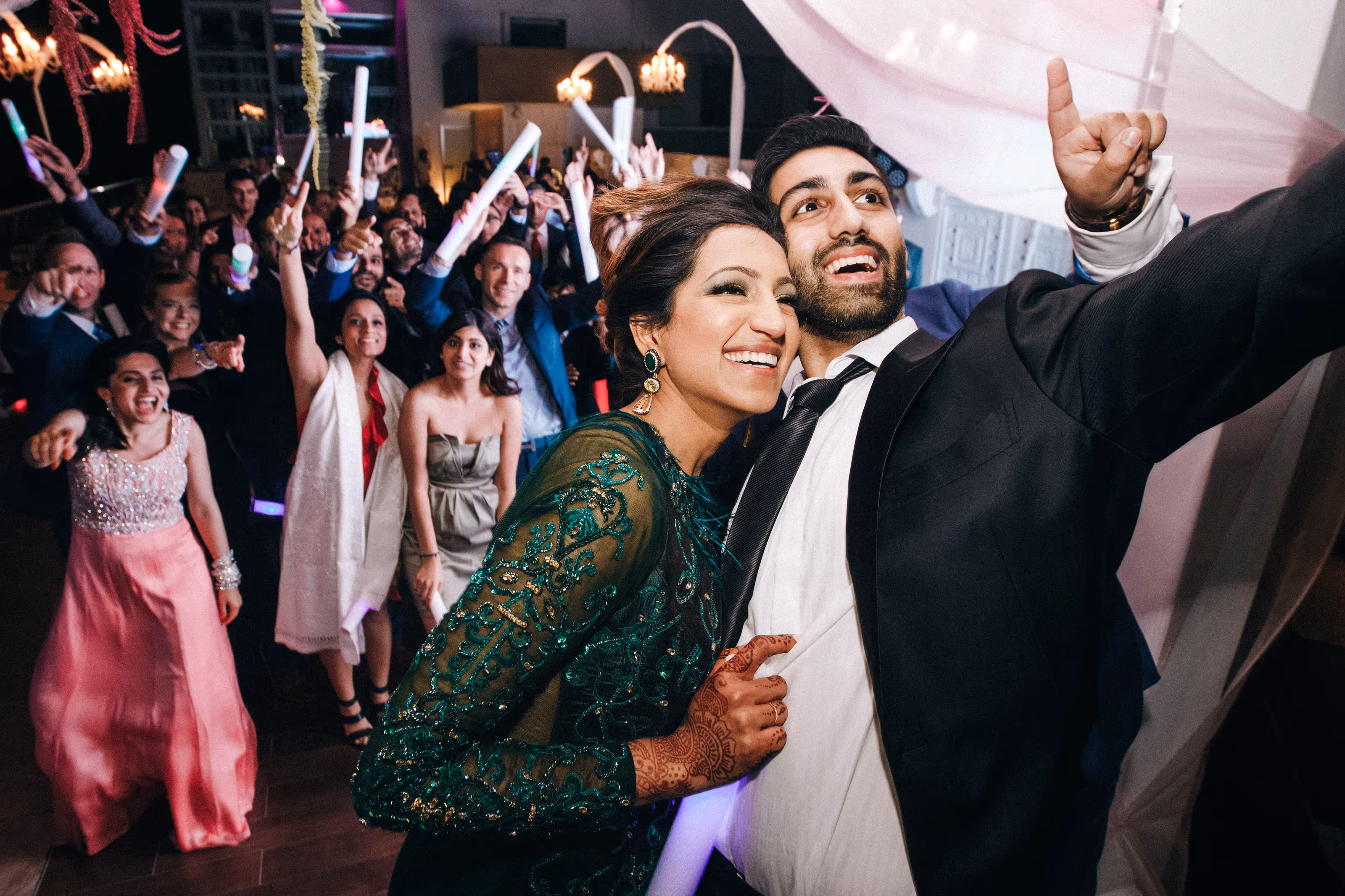 Smiling couple taking a selfie at a lively party with South Asian destination wedding guests holding glowing light sticks.