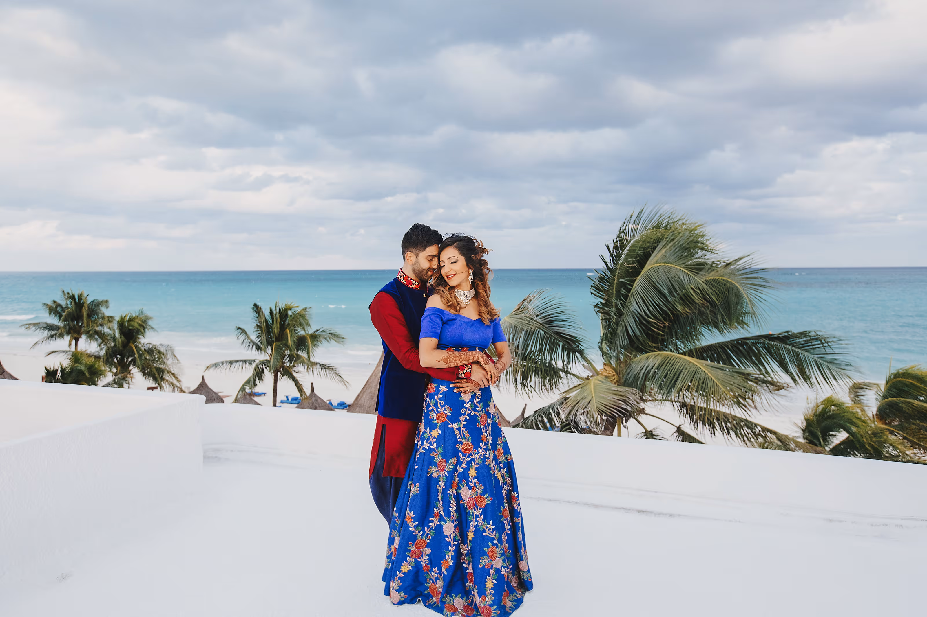 Couple in traditional South Asian attire embracing on a white rooftop by the beach with palm trees after their South Asian destination wedding.