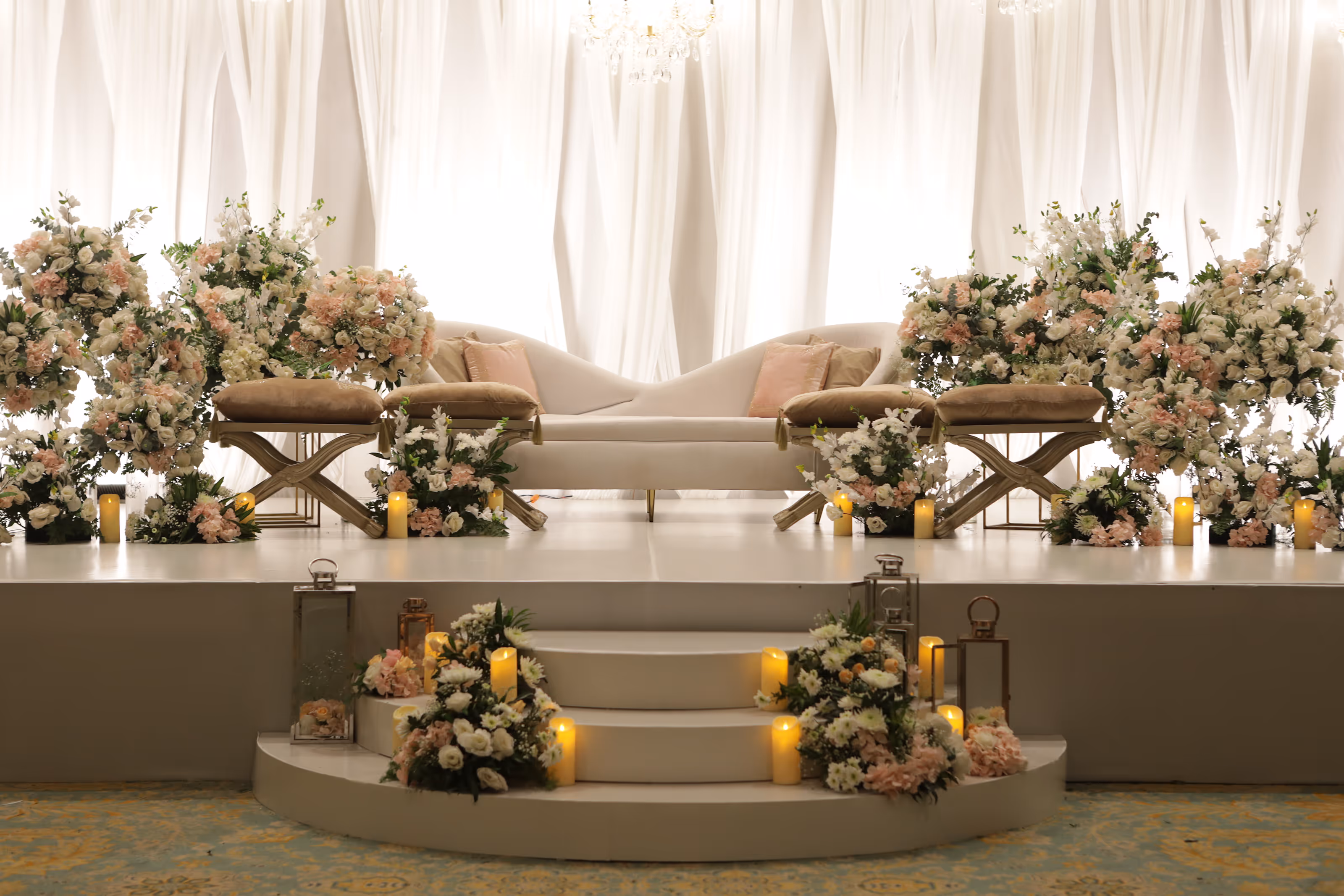 White stage with curved sofa, cushioned stools, floral arrangements, and lit candles on steps at an all-inclusive resort for a South Asian wedding.