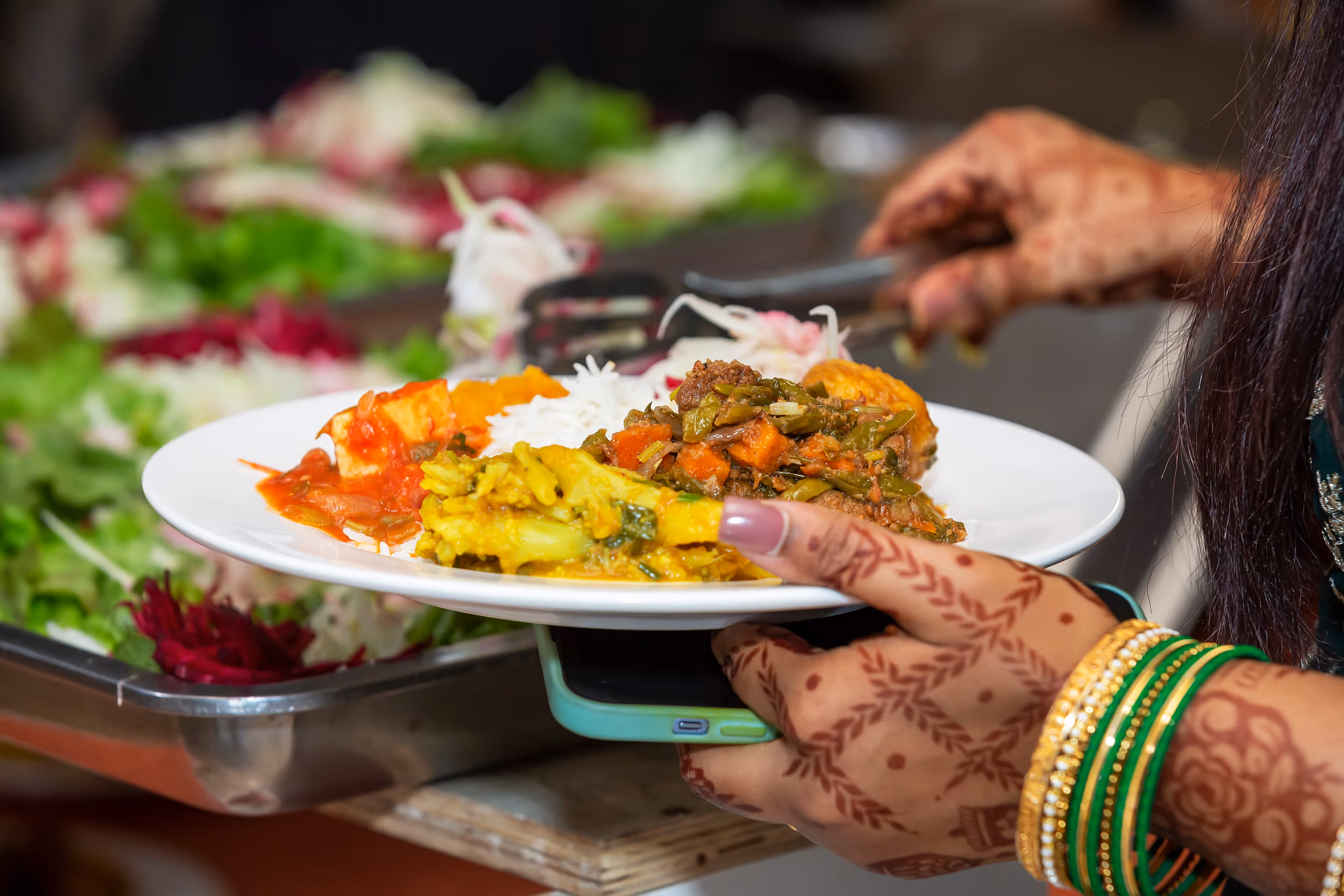Person with henna and bangles serving Indian food on a white plate from a buffet line at a South Asian destination wedding.