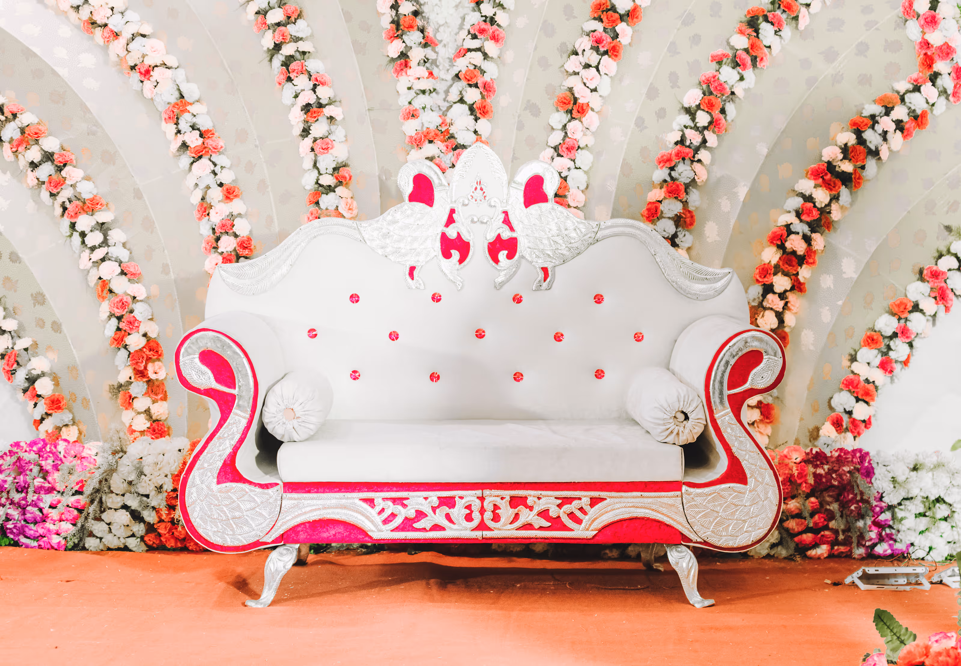 Ornate white and pink sofa with swan designs, floral backdrop, and flowers on the ground at a South Asian destination wedding.