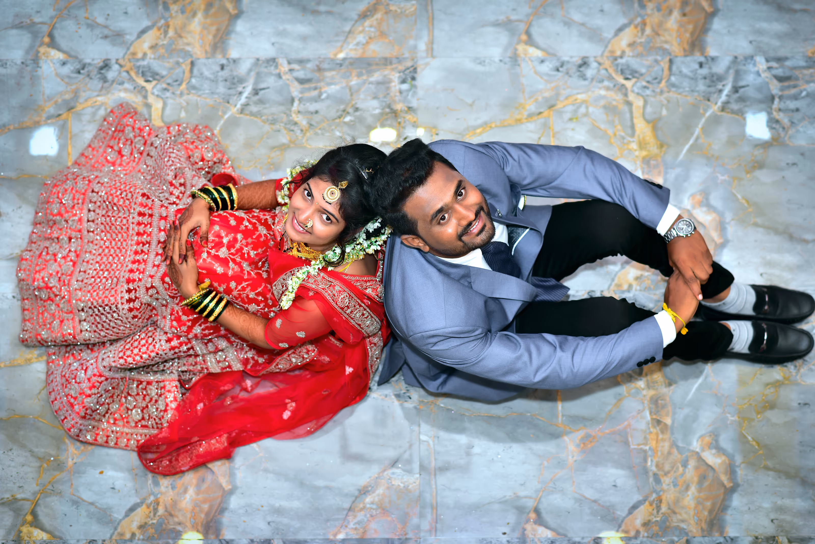 South Asian bride in red dress and groom in blue suit sitting back to back on marble floor at a destination wedding.