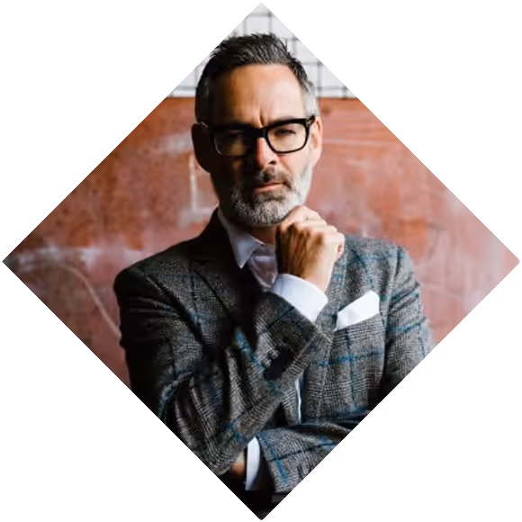 Middle-aged man with gray hair and beard, wearing glasses and a gray plaid blazer, posing thoughtfully with his hand on his chin.