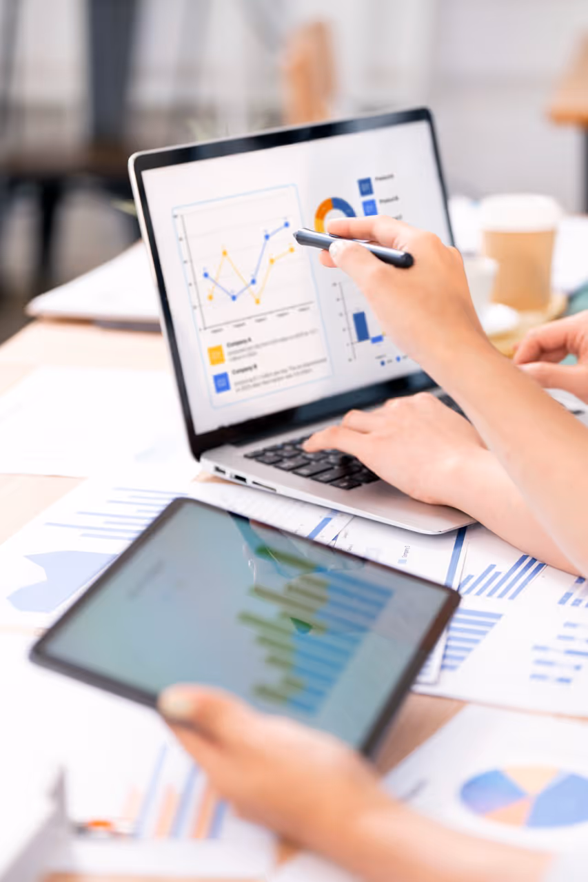 Person analyzing data charts on a laptop and a tablet, with printed graphs on the desk.