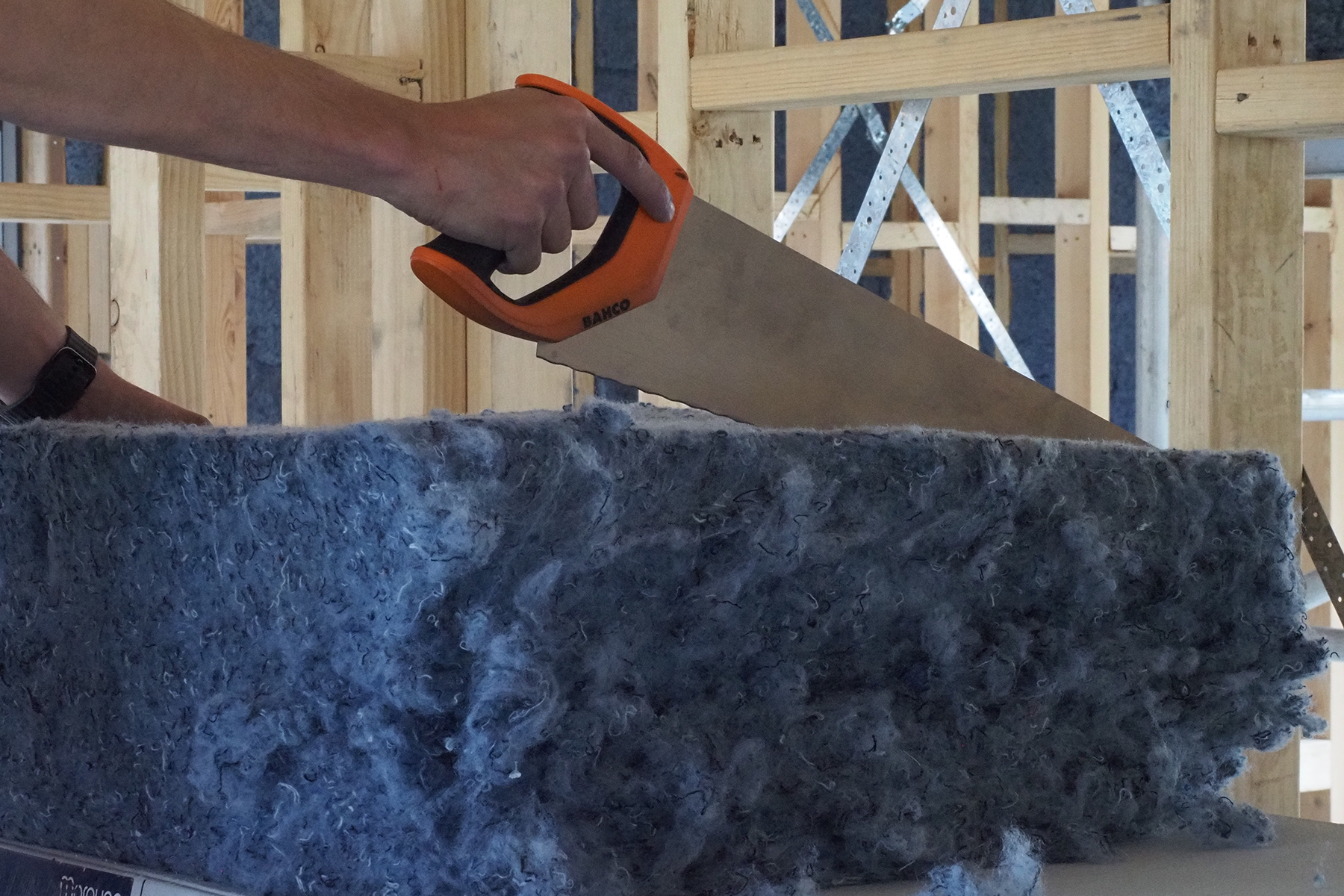 a denim blue piece of insulation is on a simple table, there is a bahco insulation saw slicing the insulation batt, just as if it were a bread knife curring bread. in the background is the framework of a house under construction